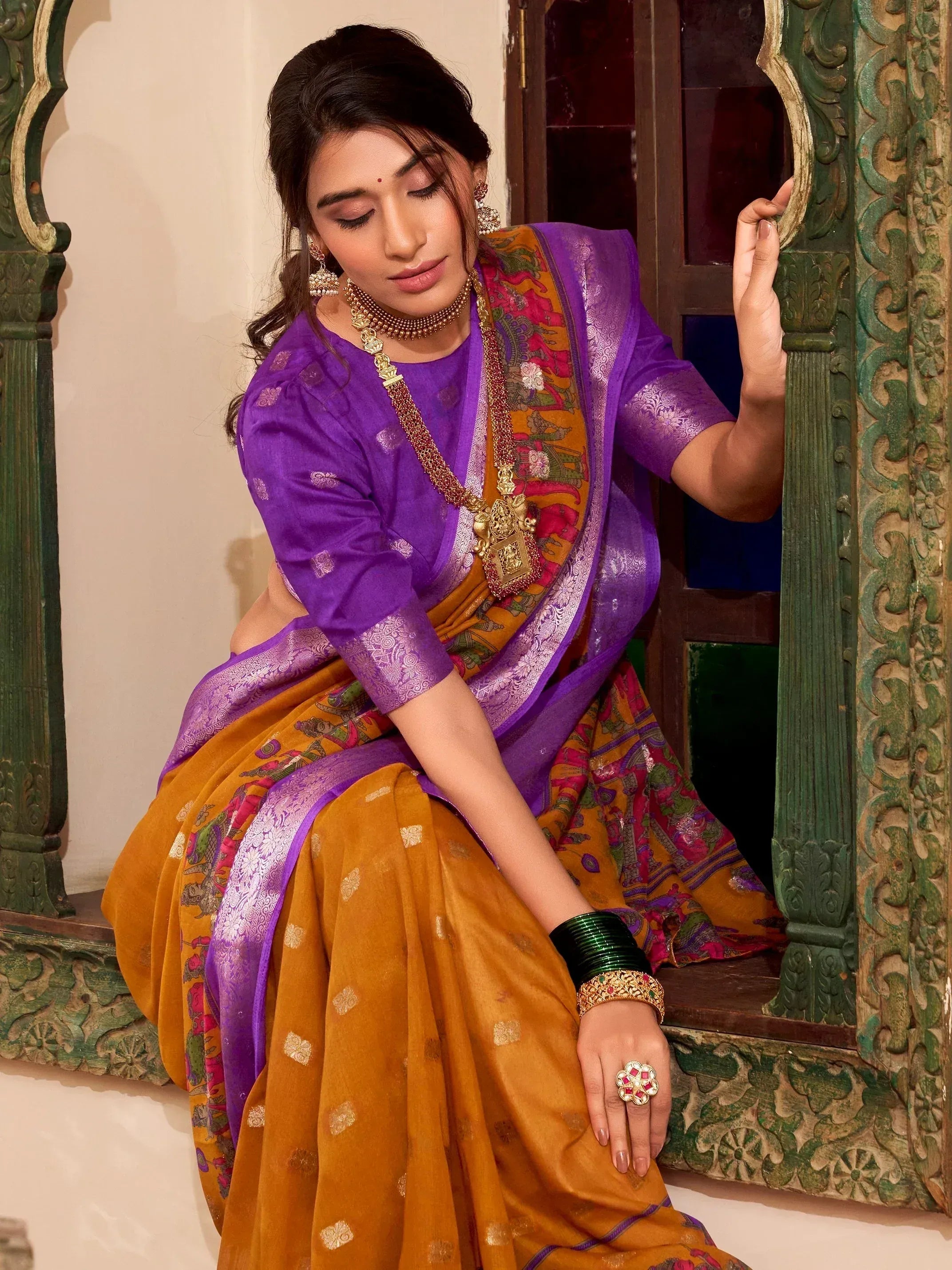 Woman in purple and mustard Indian saree with gold jewelry, seated by carved green window