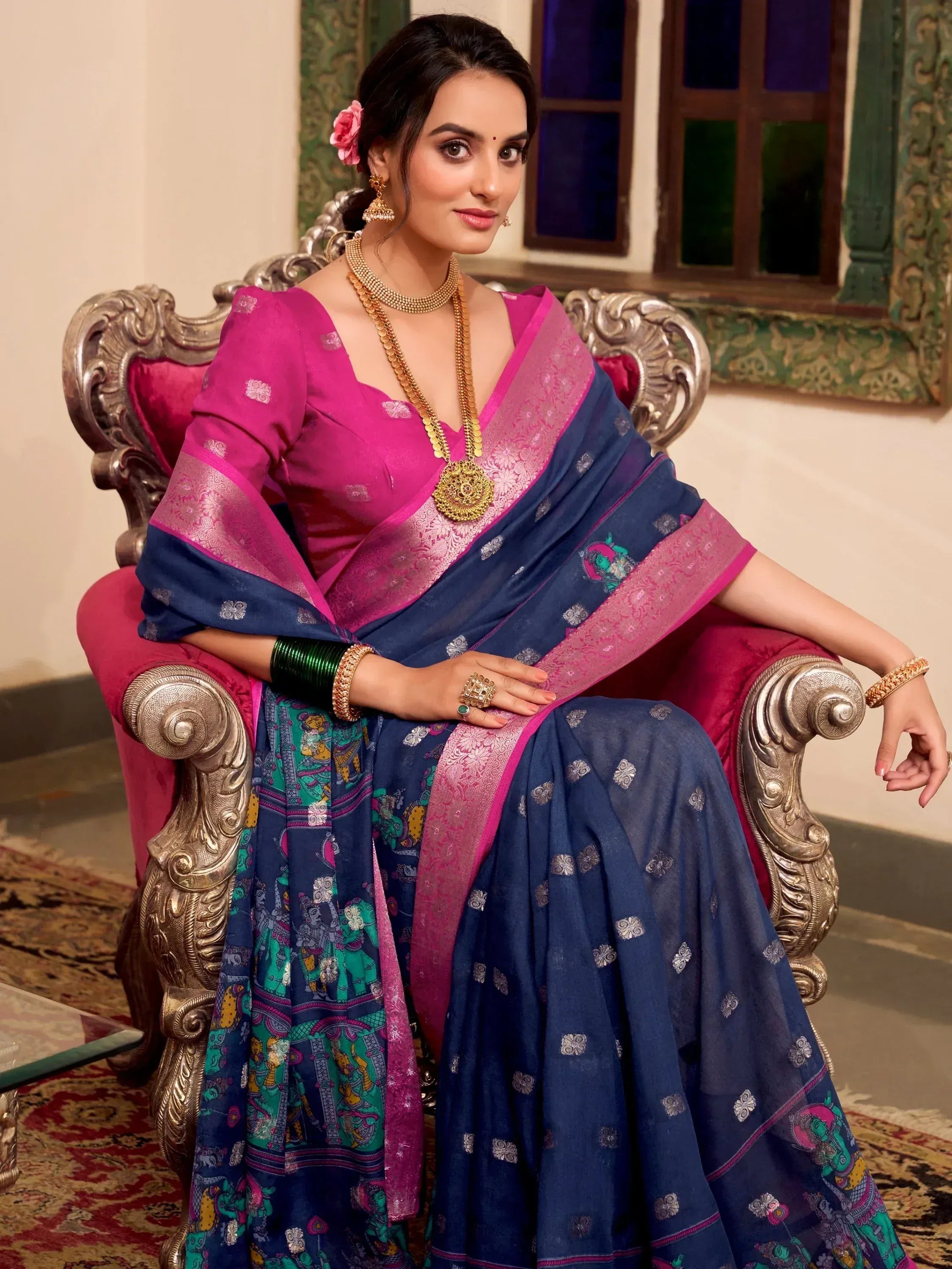 Woman in navy blue and pink ethnic saree with traditional jewelry, seated on ornate chair