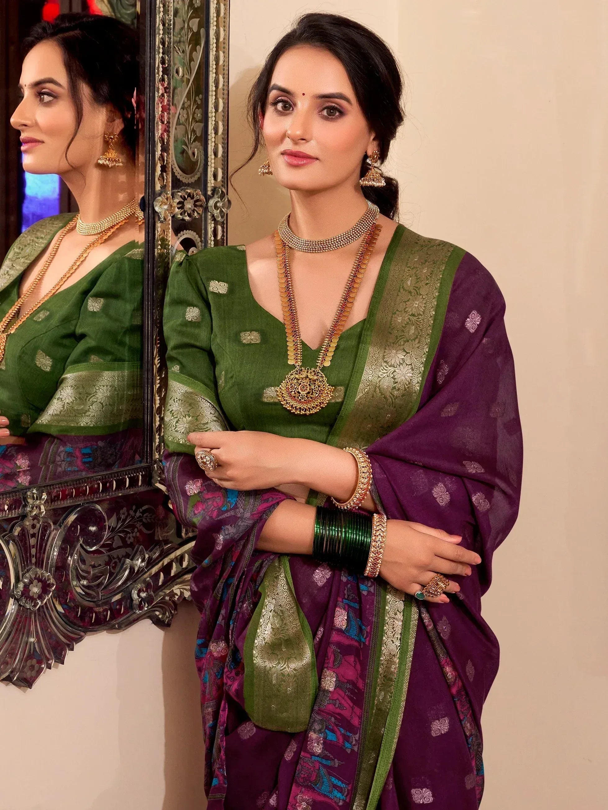 Woman in green and purple ethnic saree with gold jewelry, standing by ornate mirror