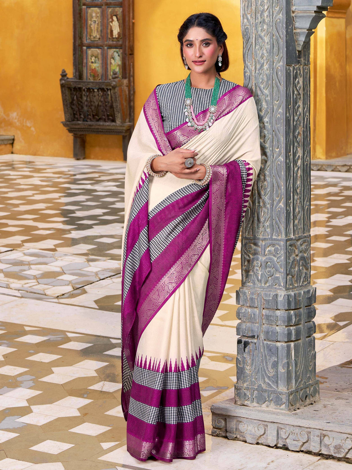 Cream and magenta ethnic saree with checkered border, worn with green jewelry against carved pillar