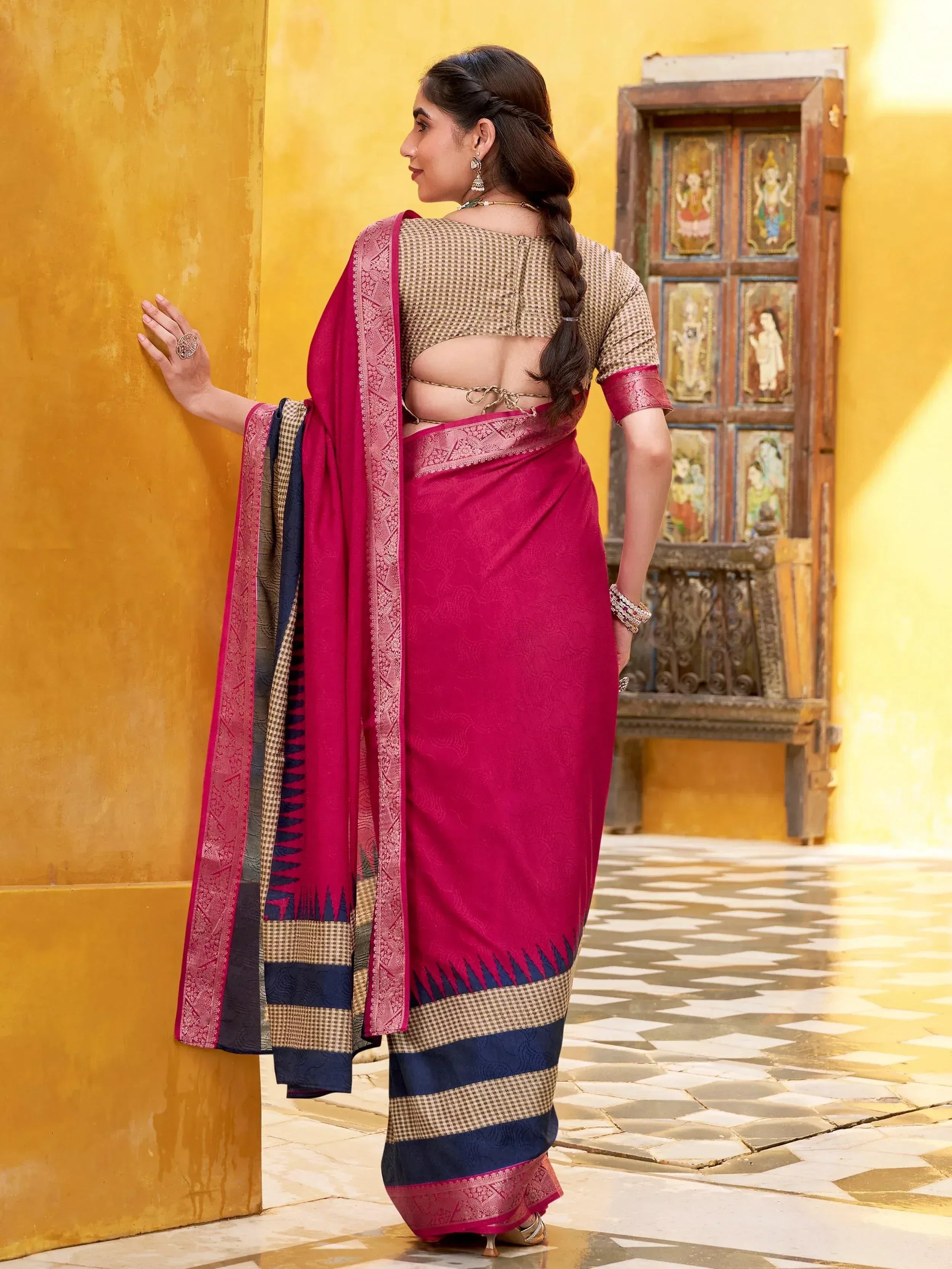 Woman in vibrant pink and navy blue saree with patterned blouse, standing by yellow wall