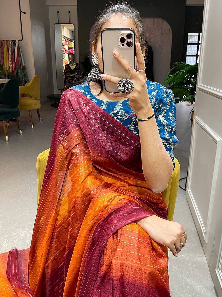 Woman in vibrant orange-red striped saree with blue embroidered blouse, taking mirror selfie in boutique