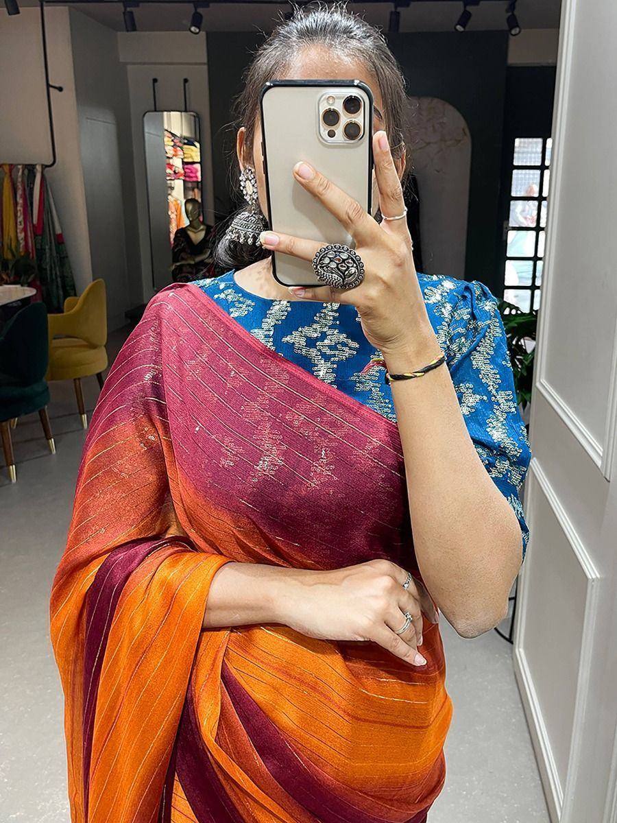 Woman in vibrant orange and maroon saree with blue embroidered blouse, taking mirror selfie in boutique