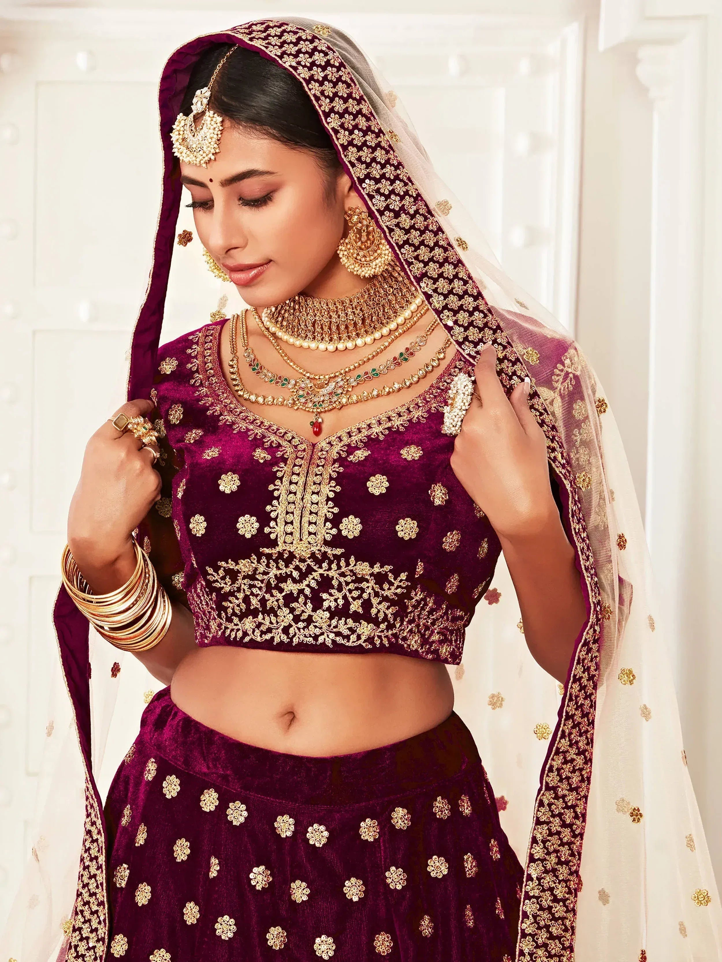 Elegant maroon velvet lehenga choli with gold embroidery, paired with traditional Indian bridal jewelry and dupatta.