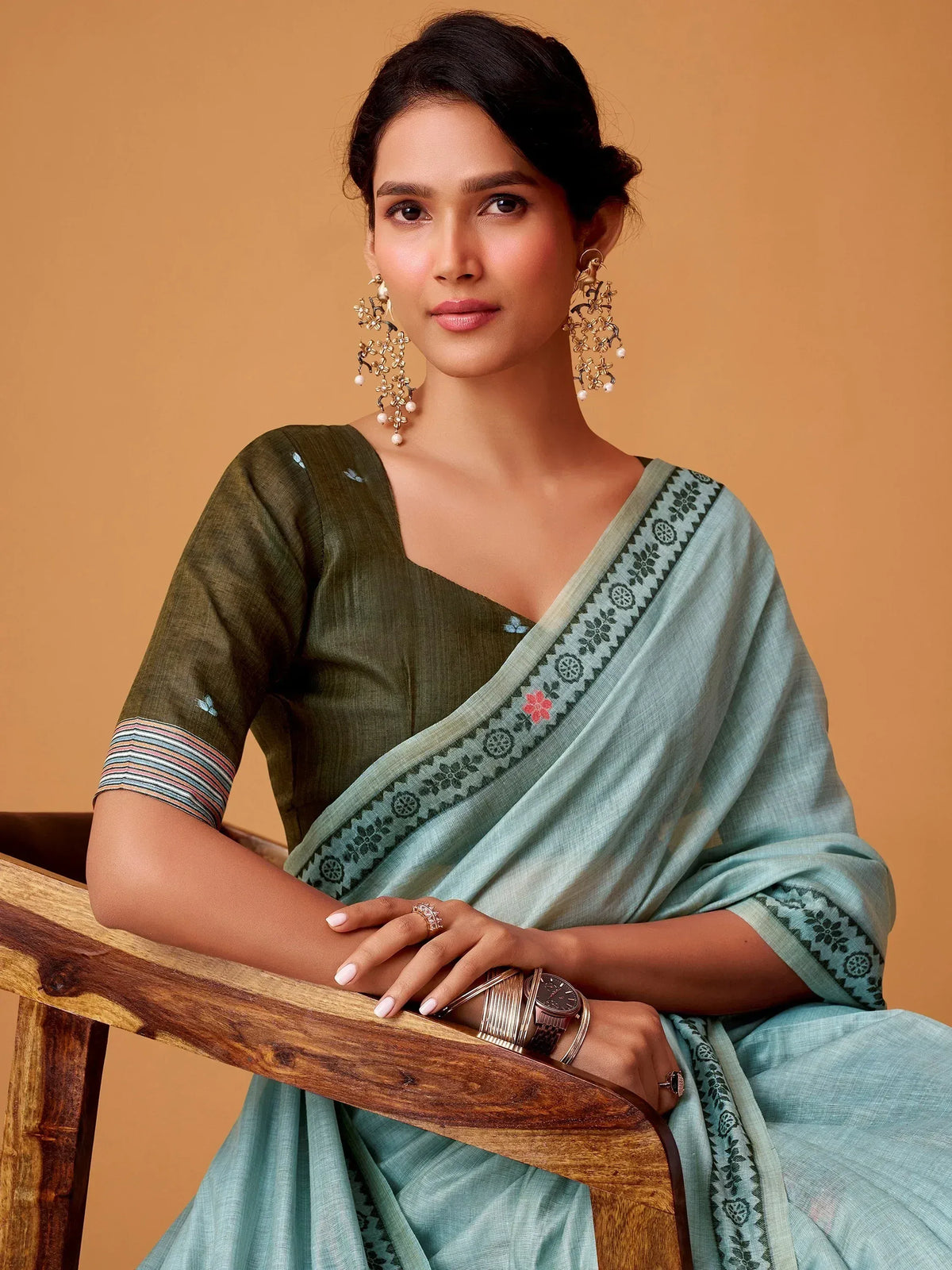 Woman in elegant light blue saree with floral border and olive green blouse, styled with statement earrings.