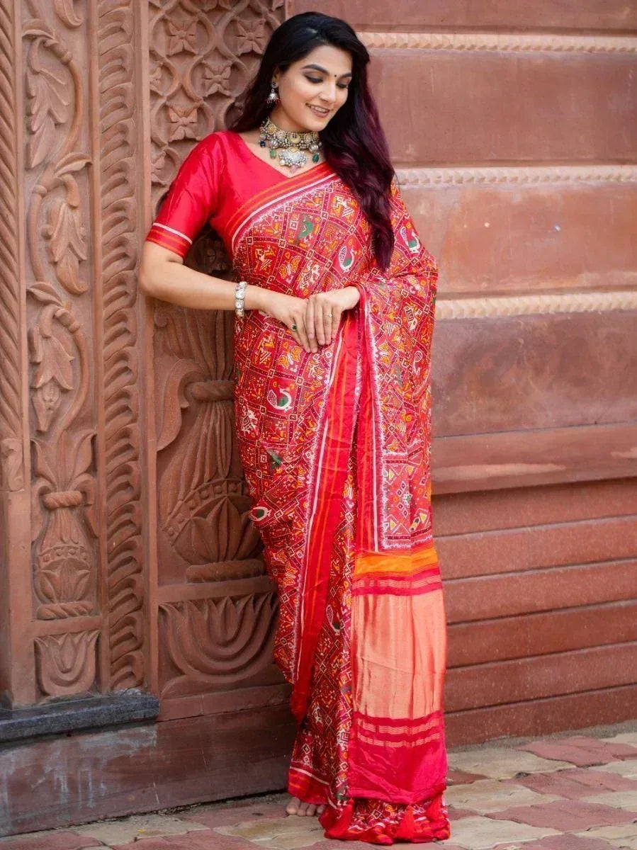 Woman in vibrant red printed saree with ethnic jewelry, standing by carved wooden door