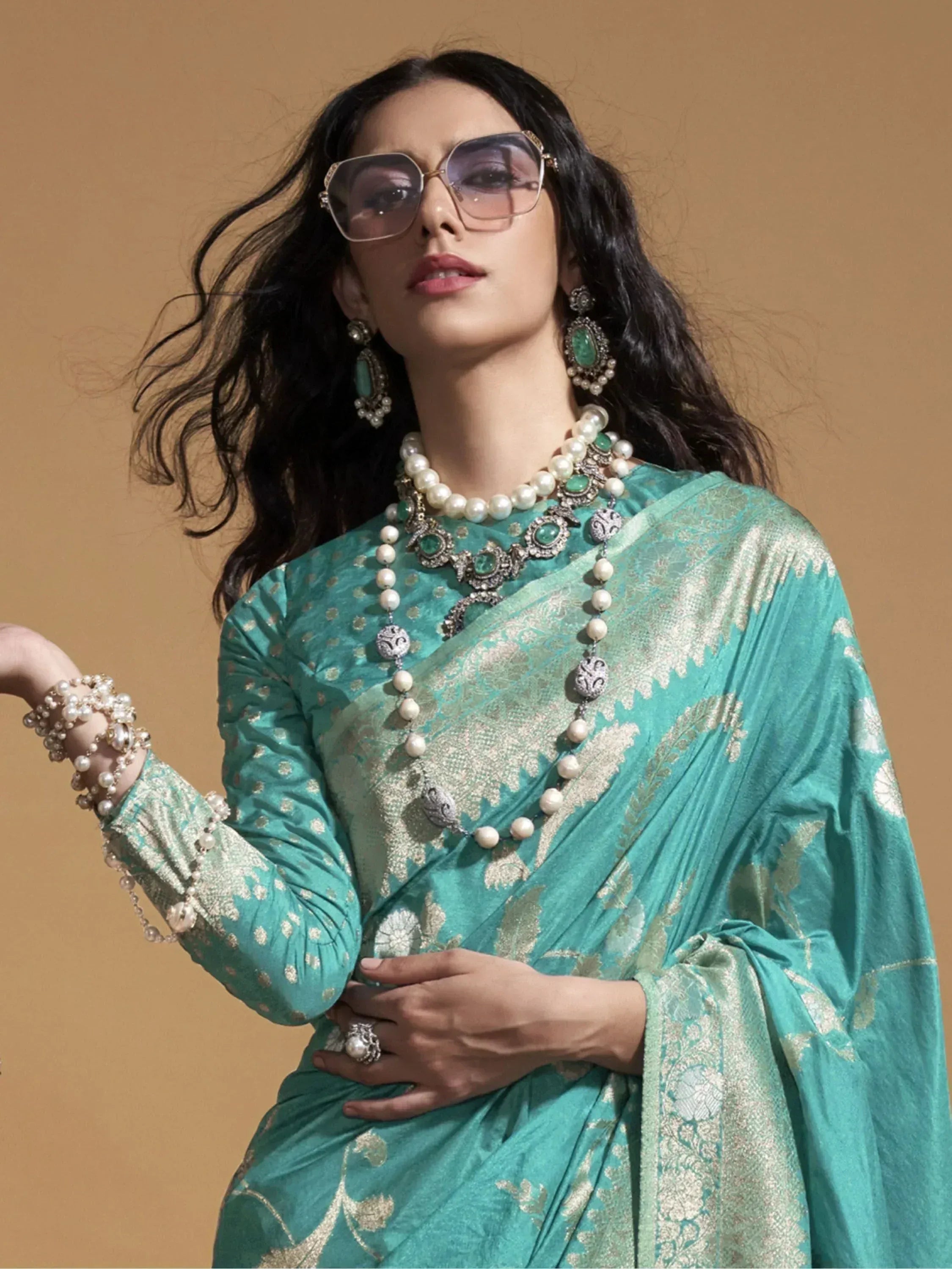 Woman in teal designer saree with gold embroidery, pearl jewelry, and statement sunglasses