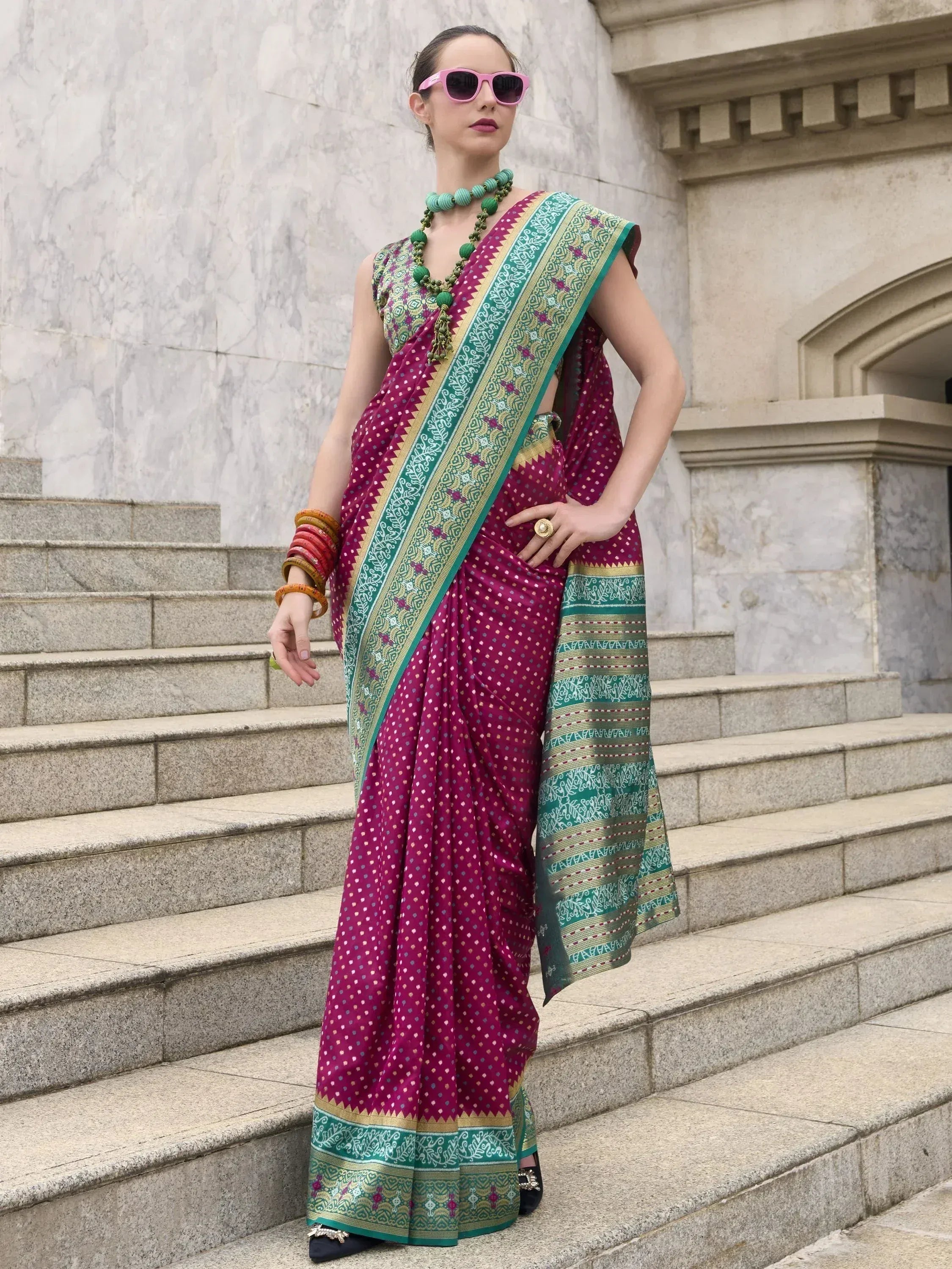 Woman in vibrant purple and green ethnic saree with intricate border, styled with sunglasses and bangles on stone steps