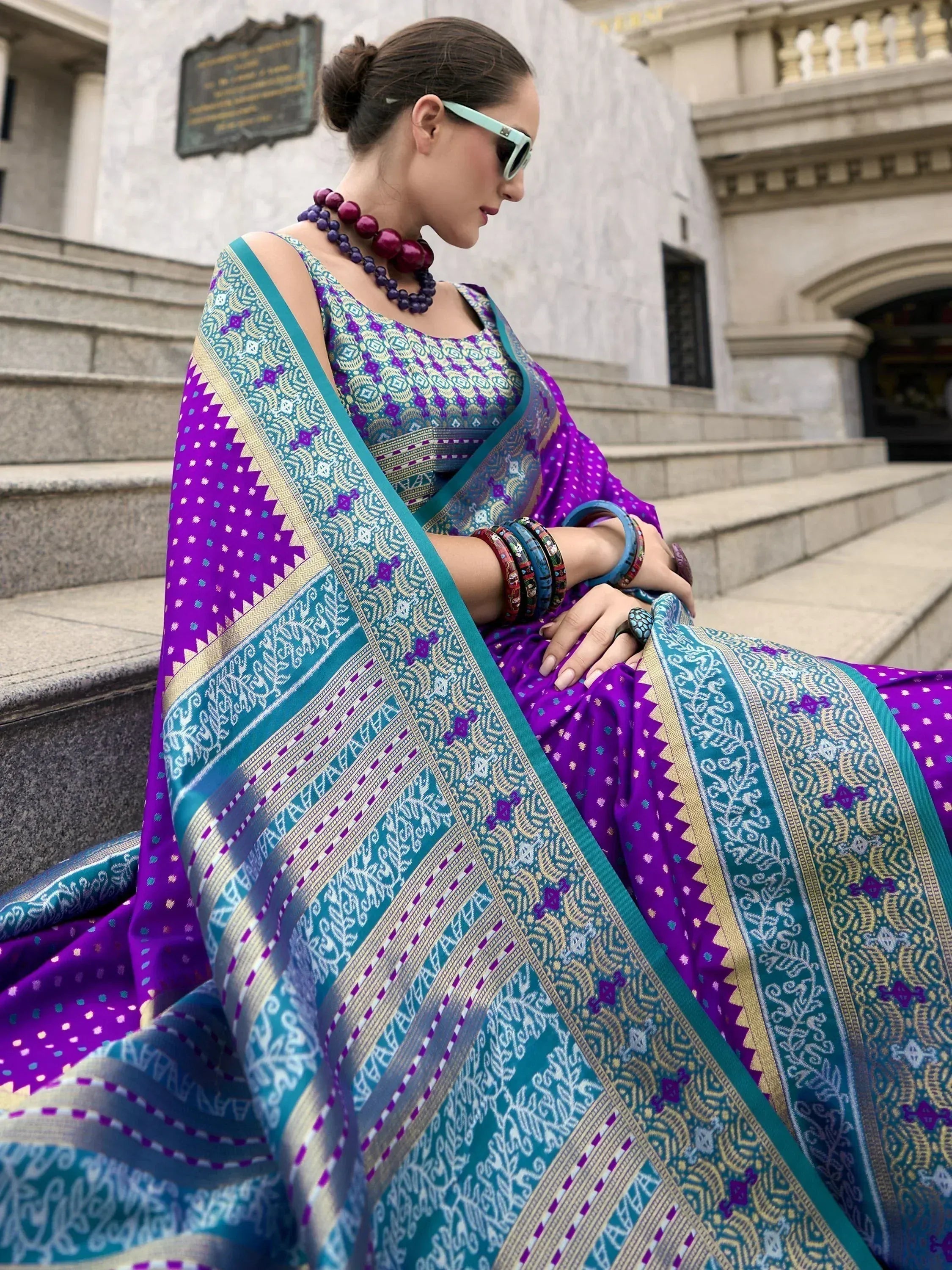 Woman in vibrant purple and teal ethnic saree with intricate patterns, styled with statement jewelry and sunglasses.