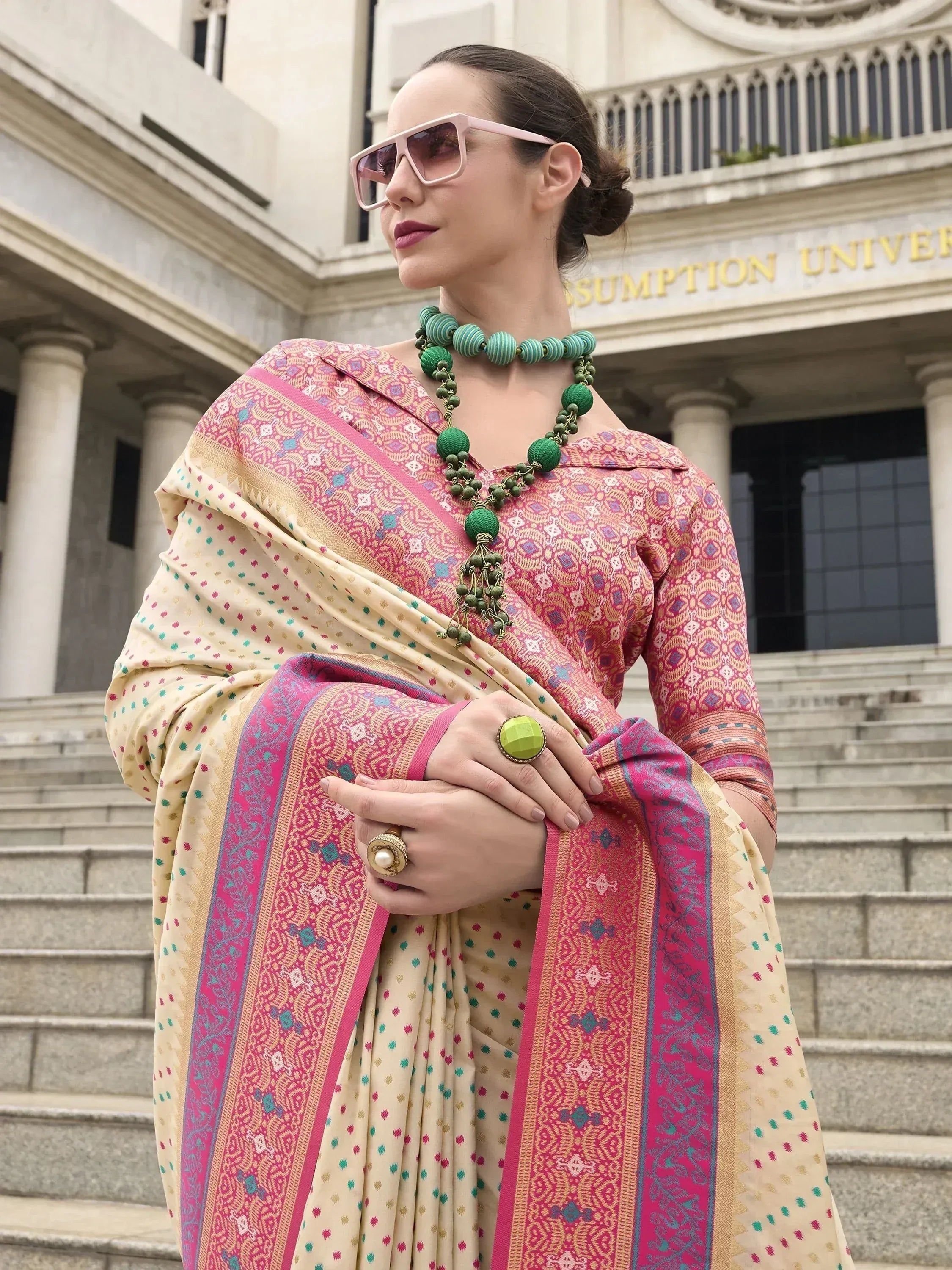 Woman in pink and cream printed saree with green jewelry, stylish sunglasses, standing on steps