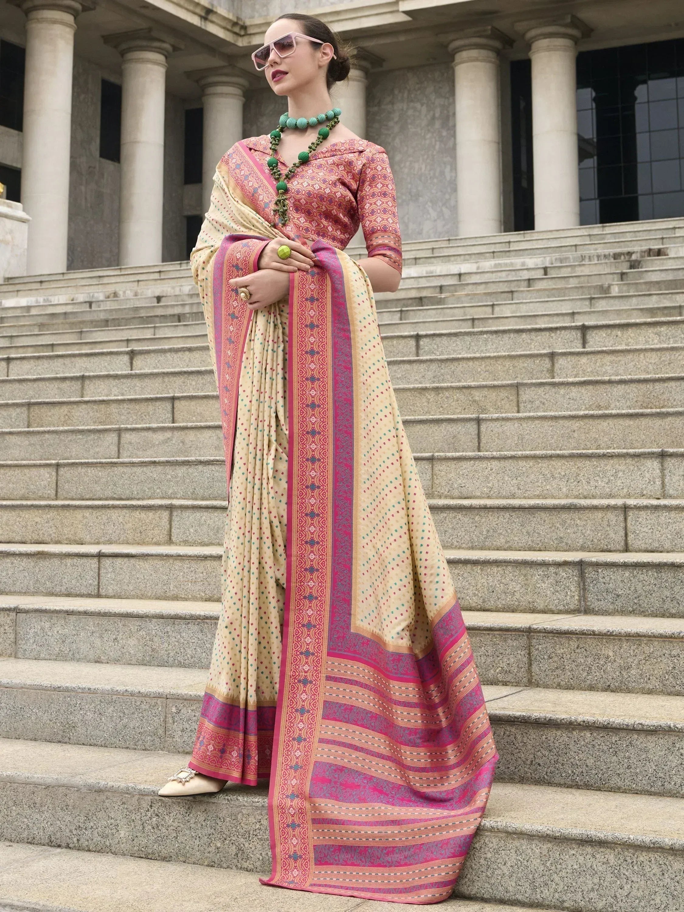 Woman in designer ethnic saree with pink border, standing on stone steps, wearing green jewelry and sunglasses