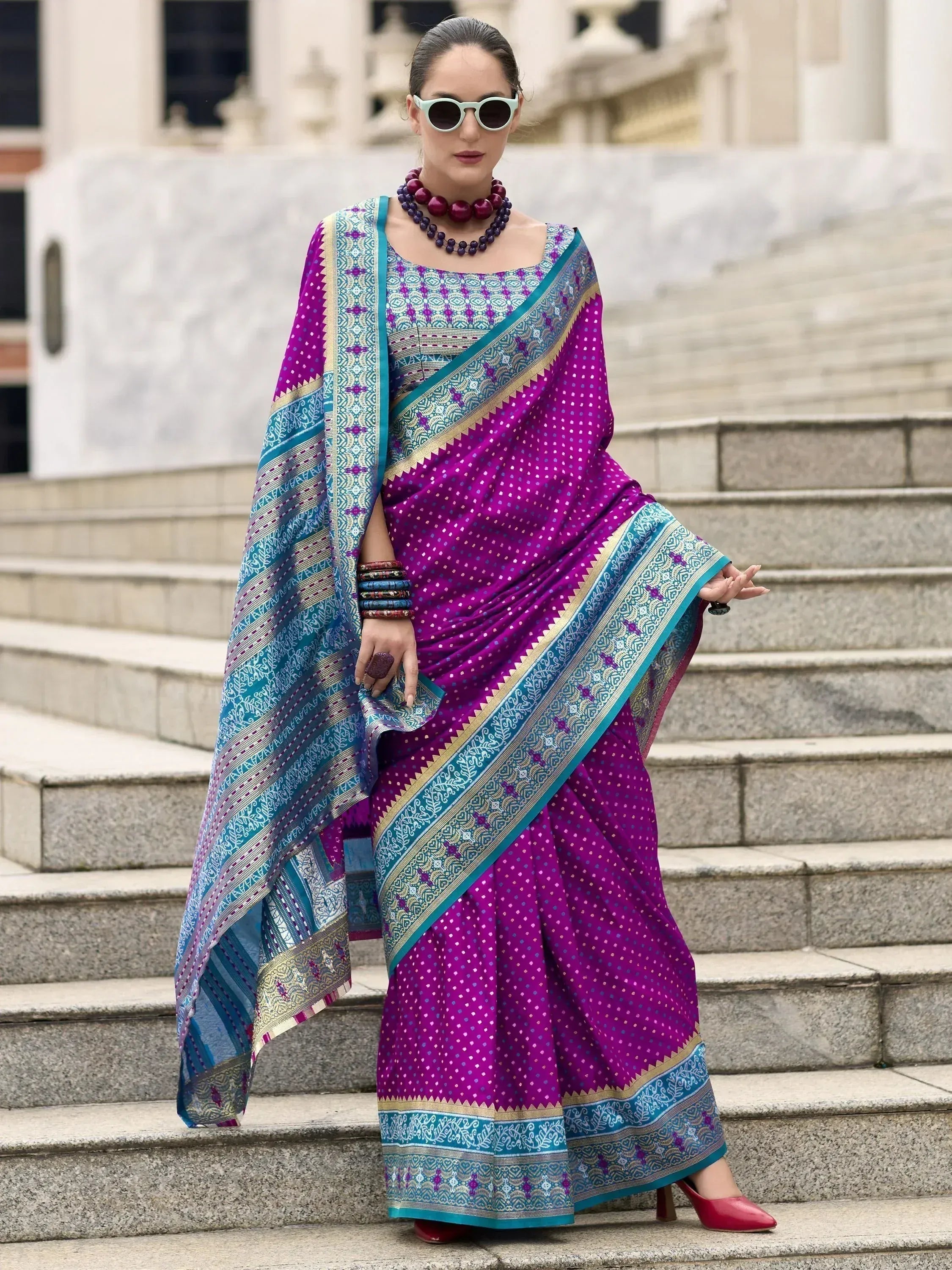 Vibrant purple and teal ethnic saree with intricate patterns, worn with statement jewelry and red heels on stone steps.