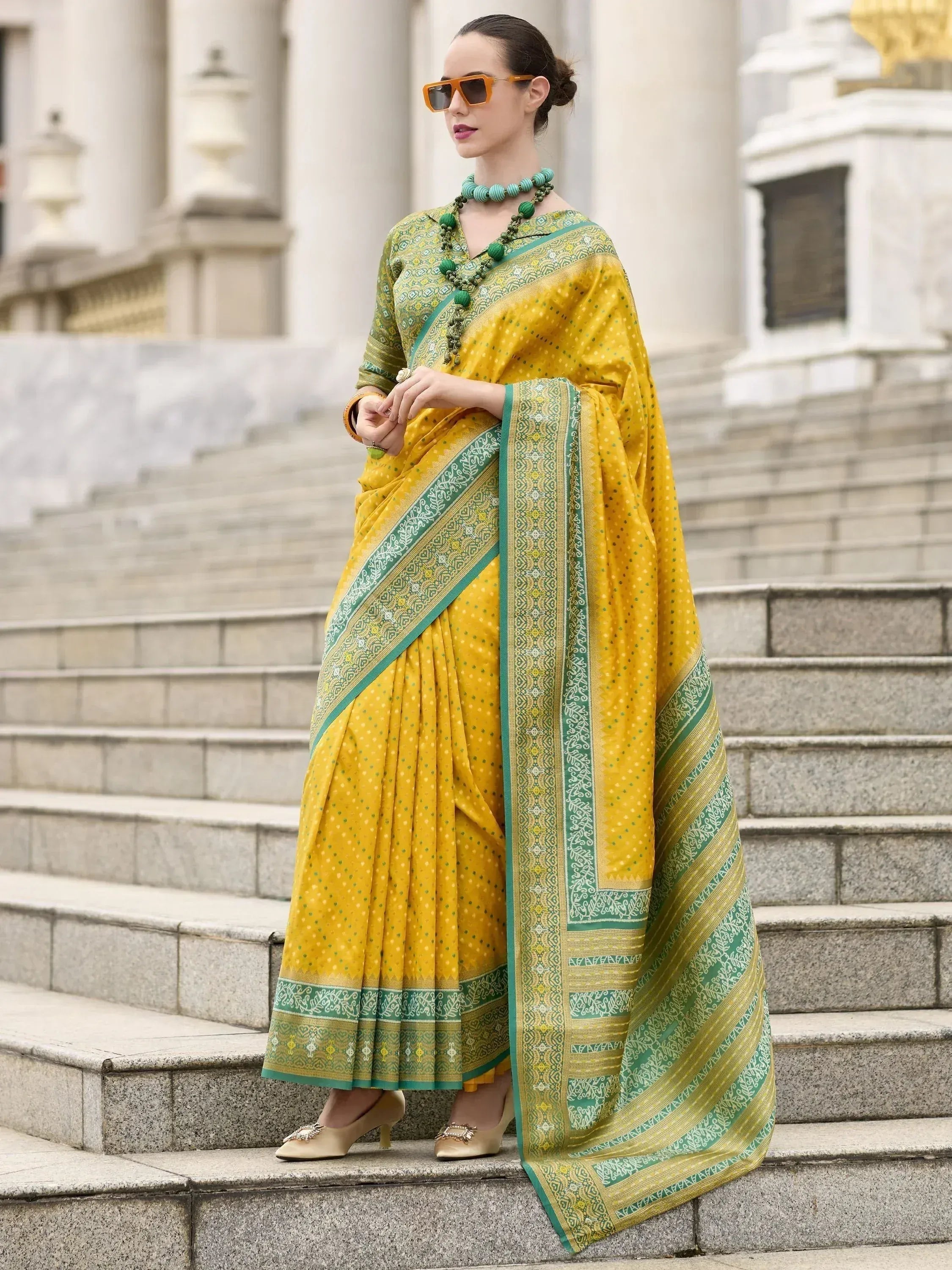 Yellow and green printed saree with intricate border, styled with sunglasses and statement jewelry on steps