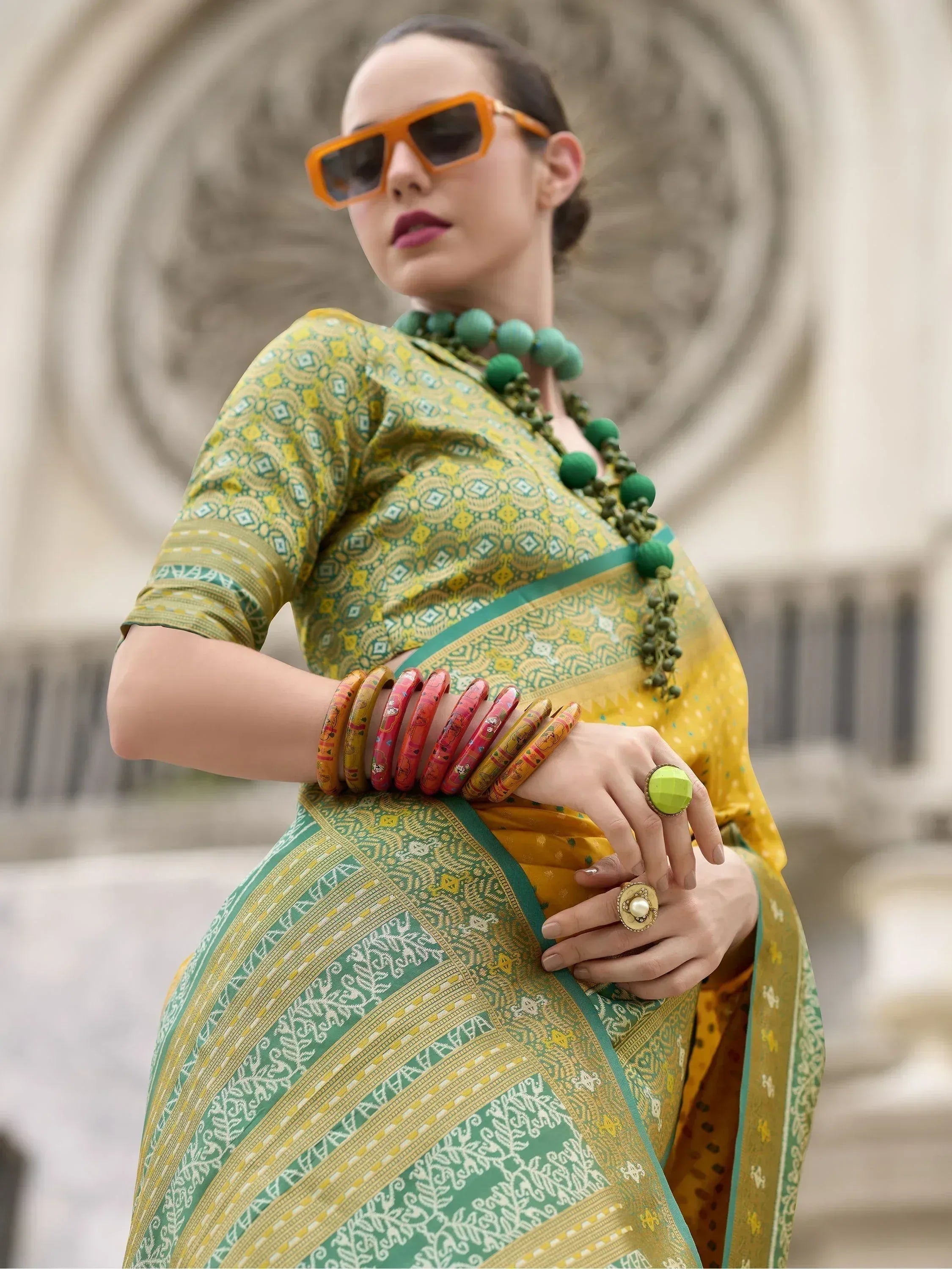 Stylish woman in vibrant yellow and green printed saree with bold accessories and orange sunglasses