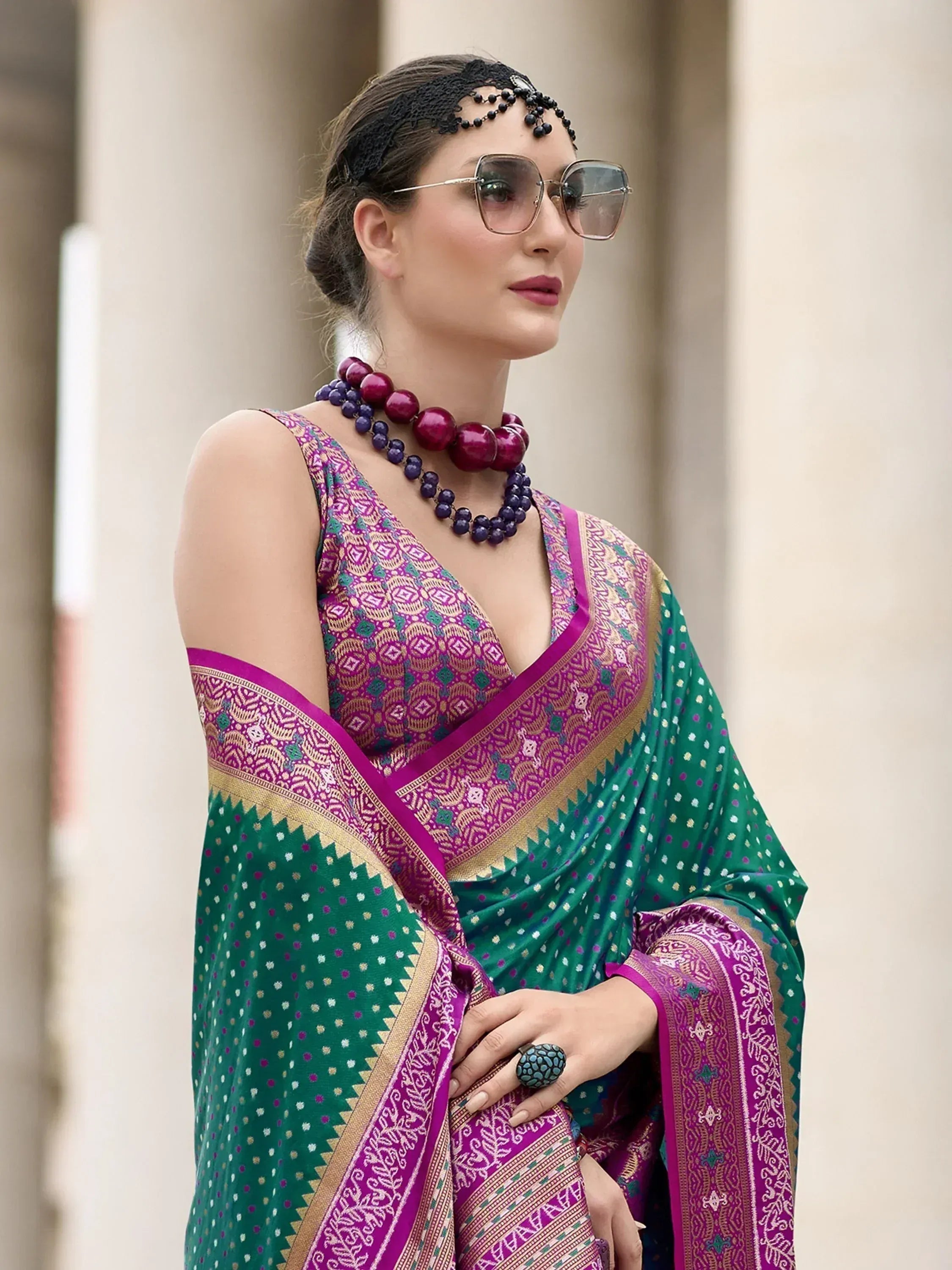 Woman in vibrant green and purple ethnic saree with intricate patterns, styled with statement jewelry and sunglasses.