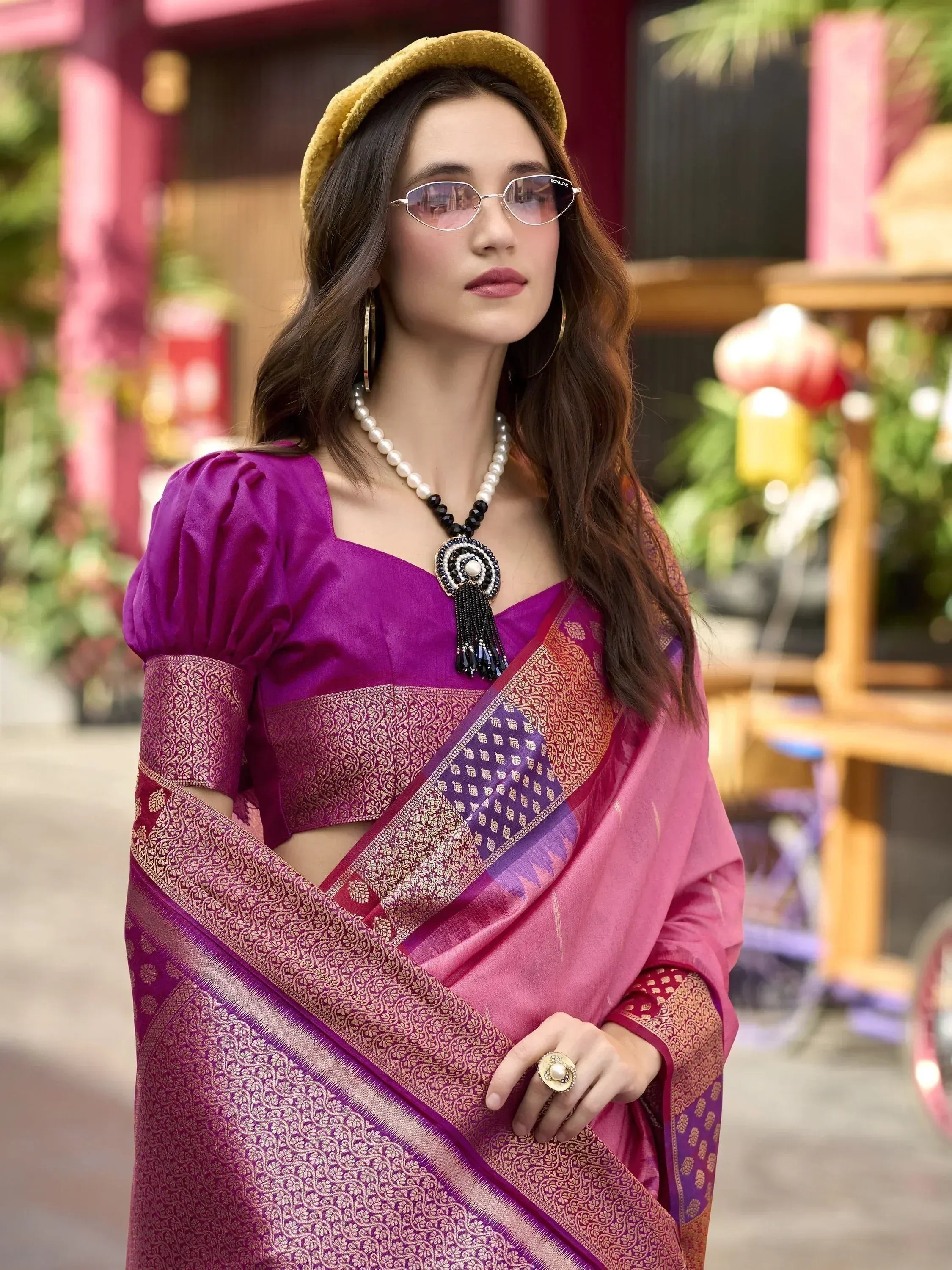Woman in purple designer saree with gold border, puff sleeves, and pearl necklace, styled with yellow hat and sunglasses.