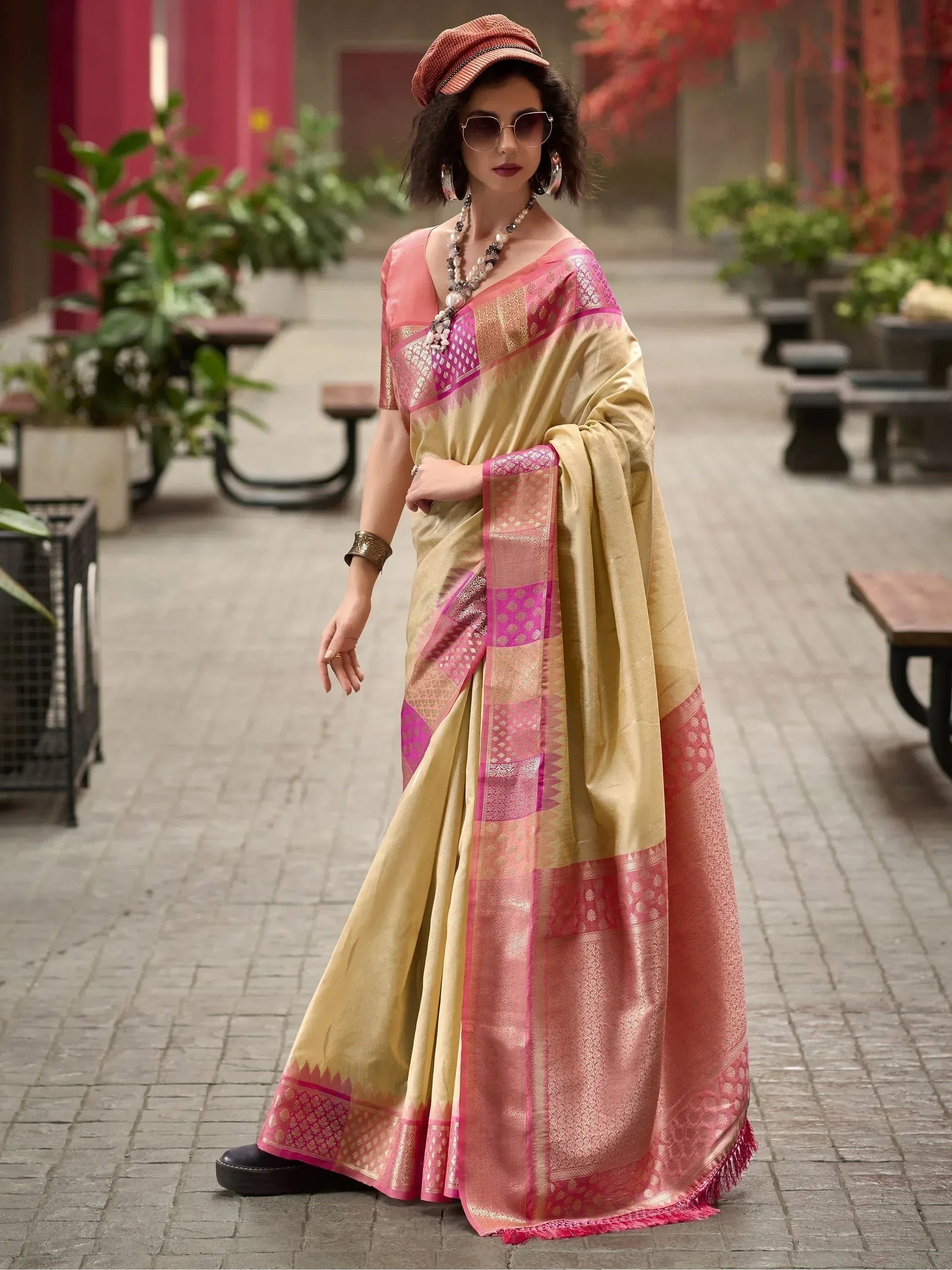 Woman in elegant gold and pink designer saree with intricate patterns, styled with cap and sunglasses for a modern ethnic look.