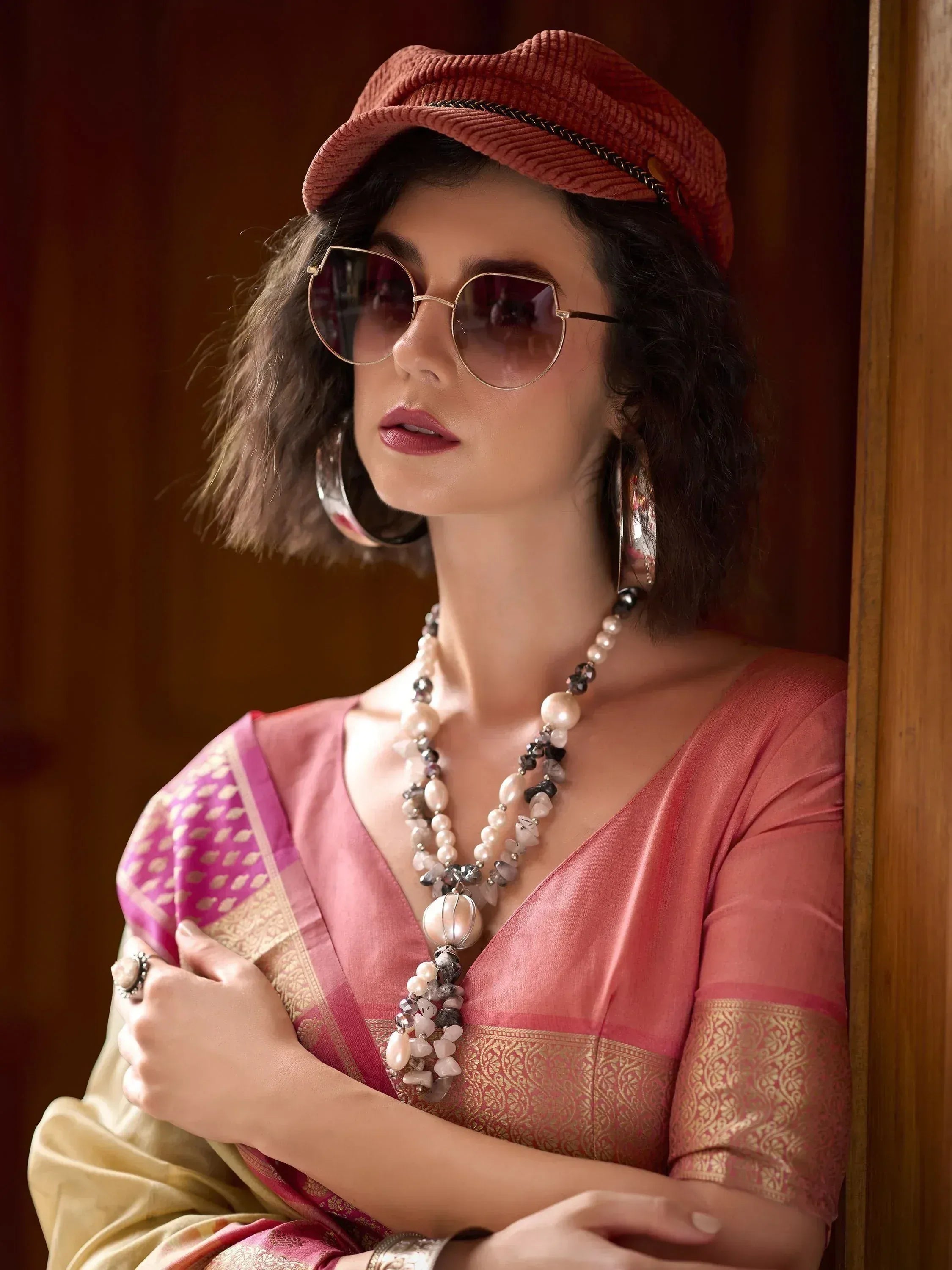 Woman in pink ethnic saree with gold border, stylish hat and sunglasses, accessorized with statement jewelry