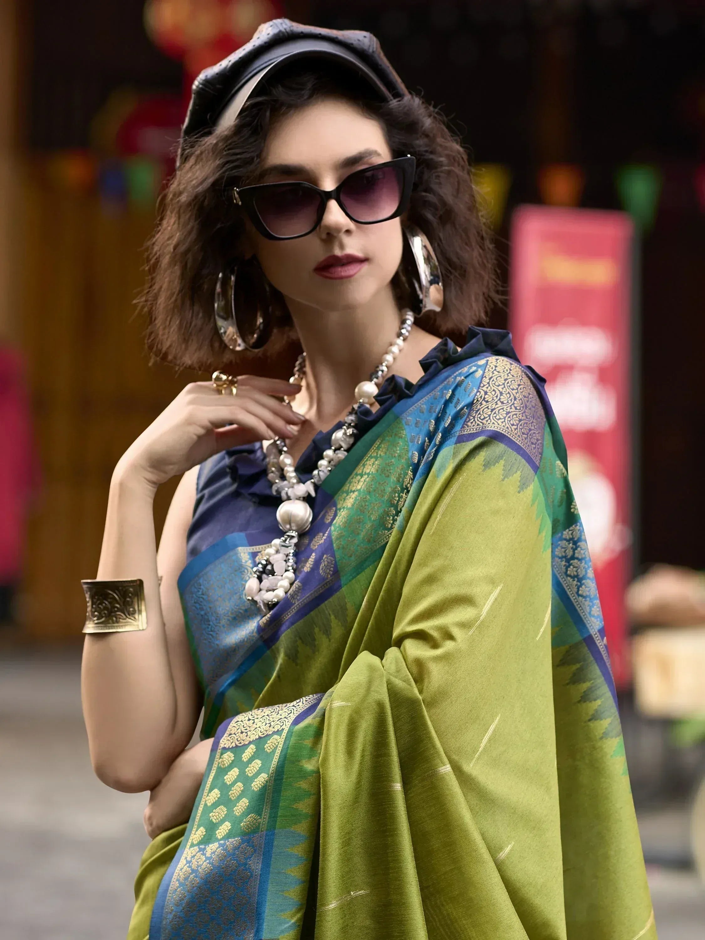 Stylish woman in green and blue ethnic saree with gold border, sunglasses, and statement jewelry.