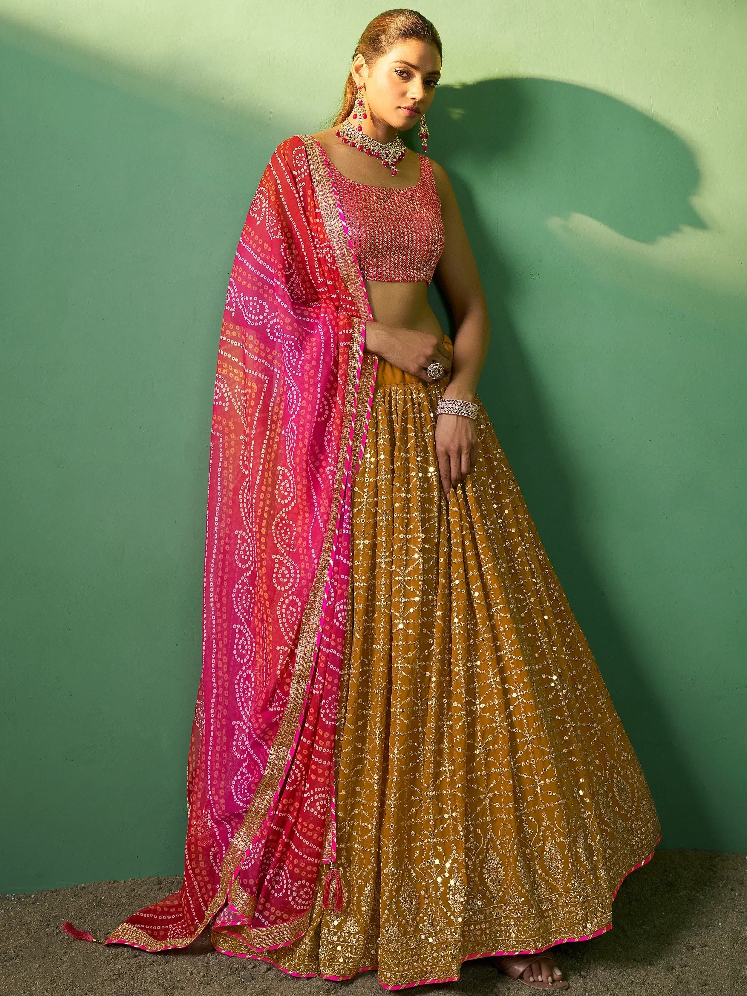 Woman in designer lehenga choli with pink dupatta, gold sequin skirt, and ethnic jewelry against green wall