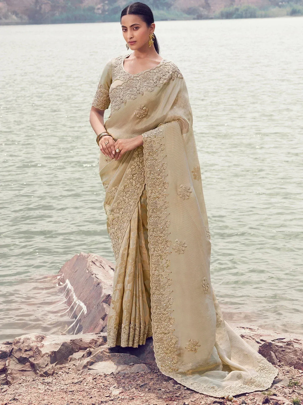 Beige embroidered saree with gold detailing, worn by woman near water, elegant Indian ethnic wear