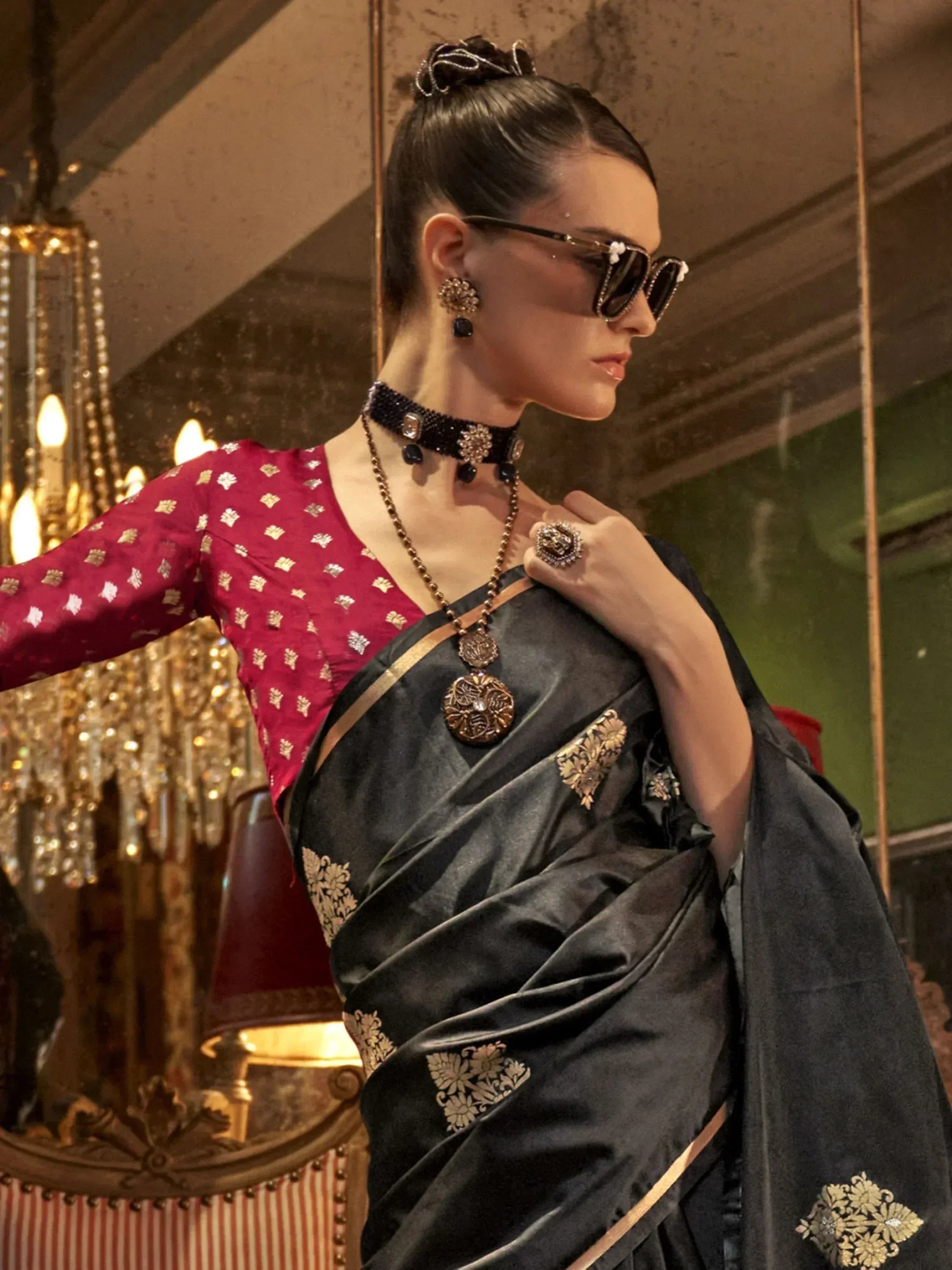 Elegant woman in black saree with gold embroidery and maroon blouse, styled with statement jewelry and sunglasses.