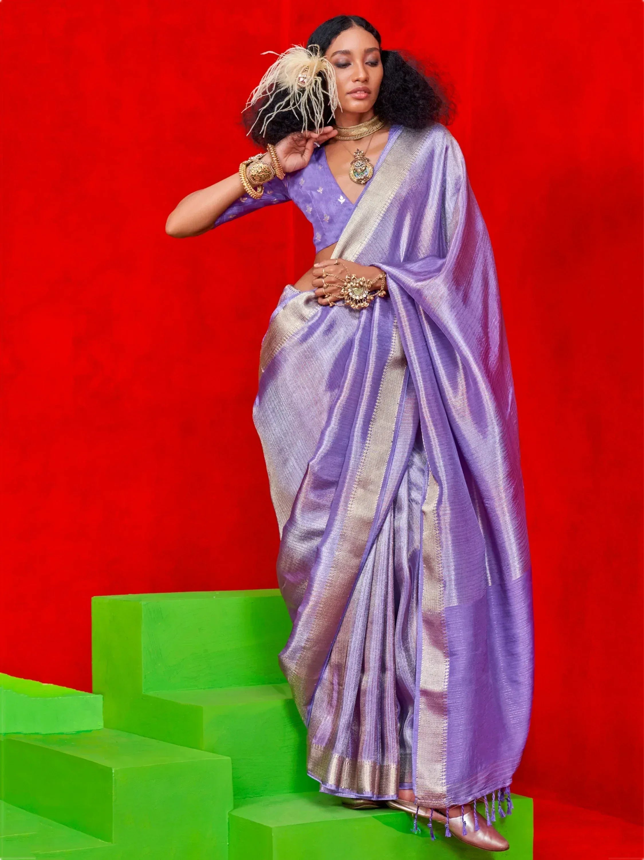 Purple designer saree with gold border, styled with traditional jewelry against red backdrop