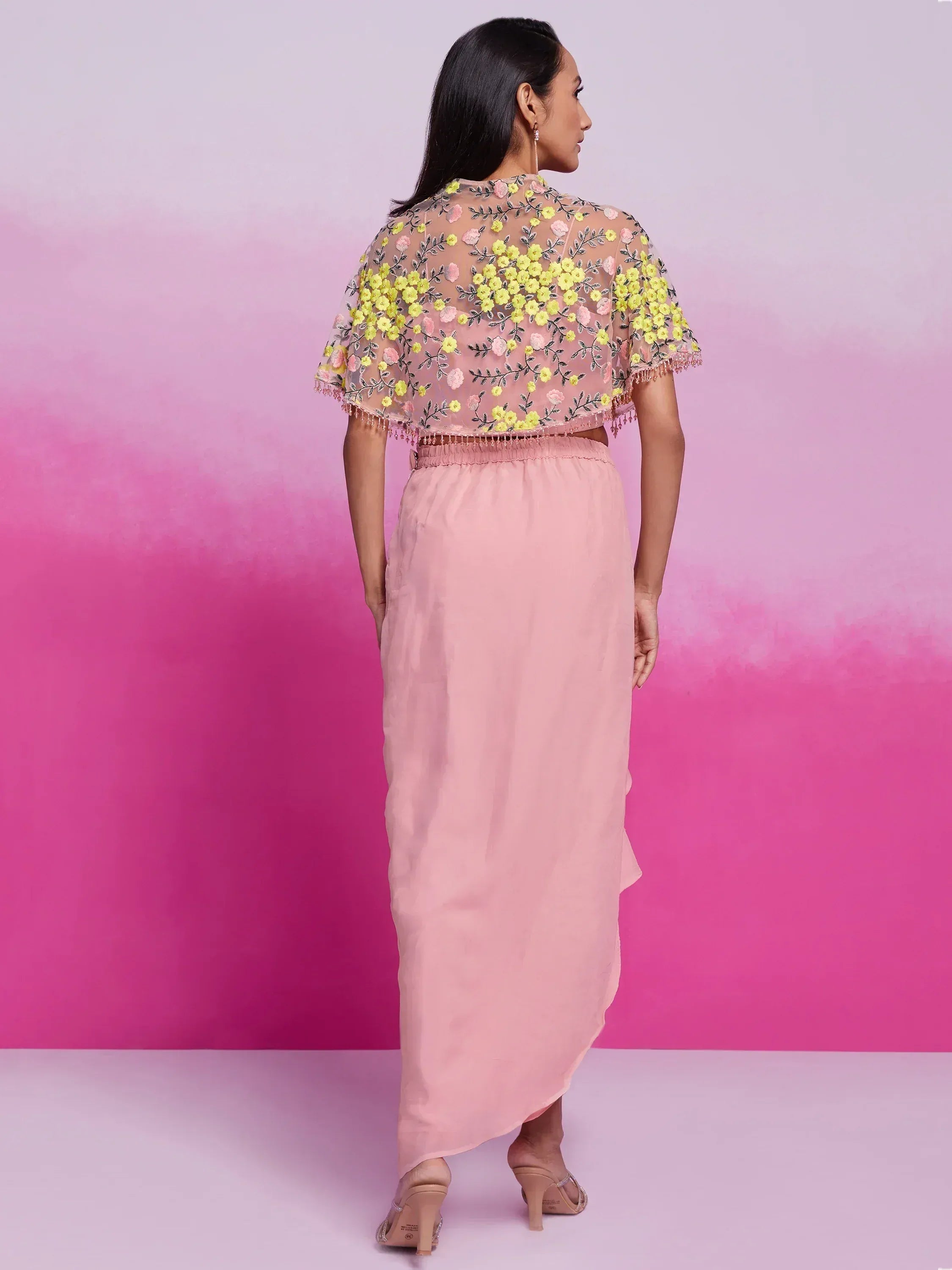 Woman in light pink saree with yellow floral embroidered cape, standing on pink background
