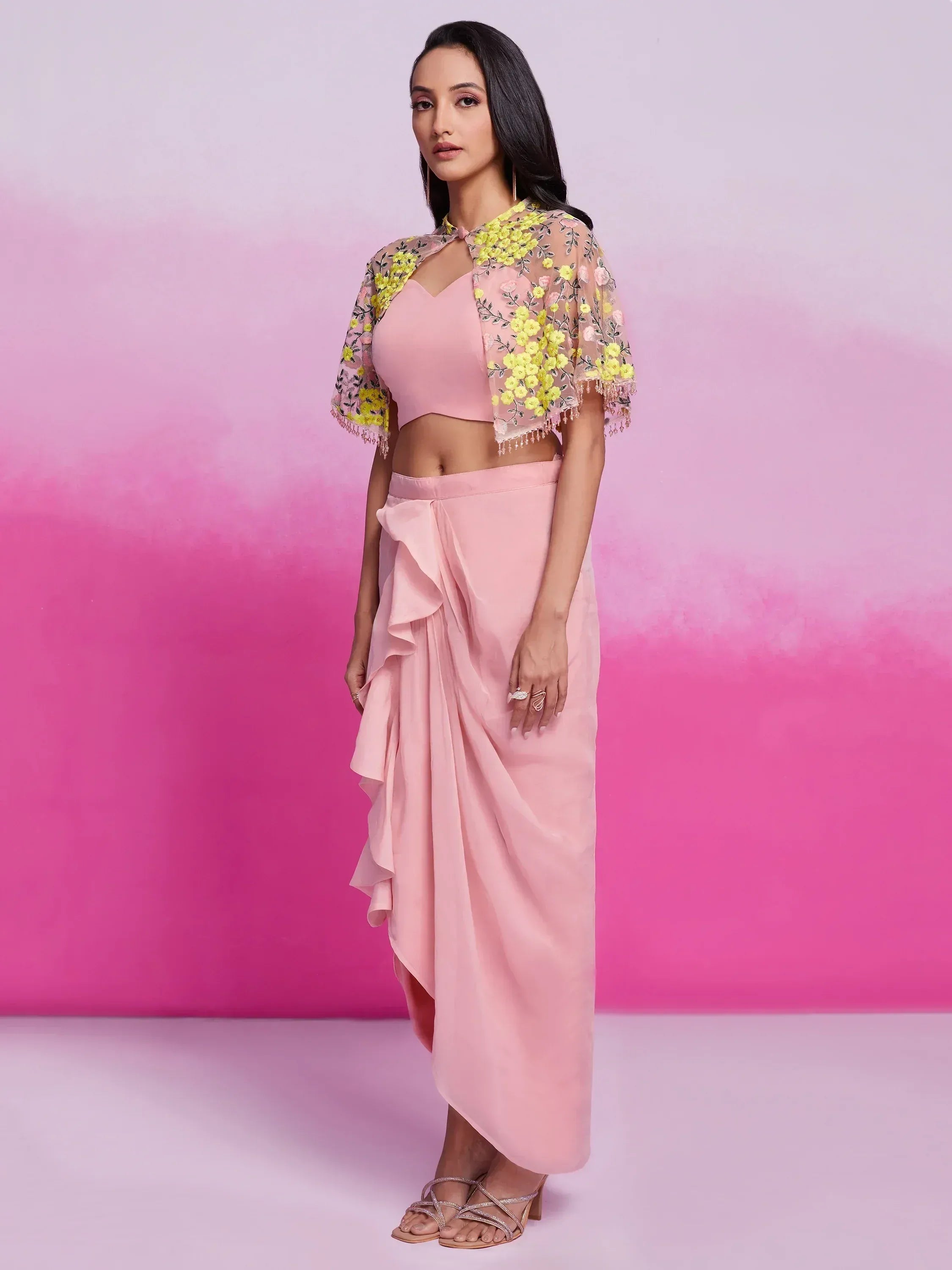 Woman in a pastel pink saree with draped ruffle skirt and sheer yellow floral cape, posing indoors