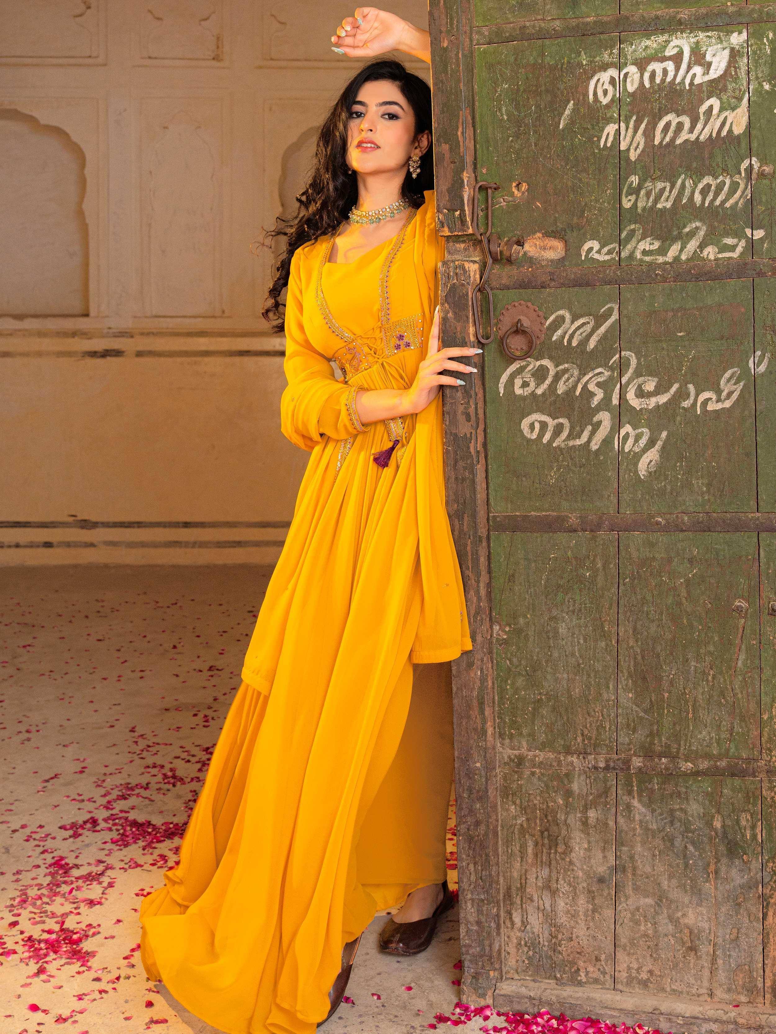 Woman in vibrant yellow ethnic gown with embroidery, leaning against rustic door, rose petals scattered