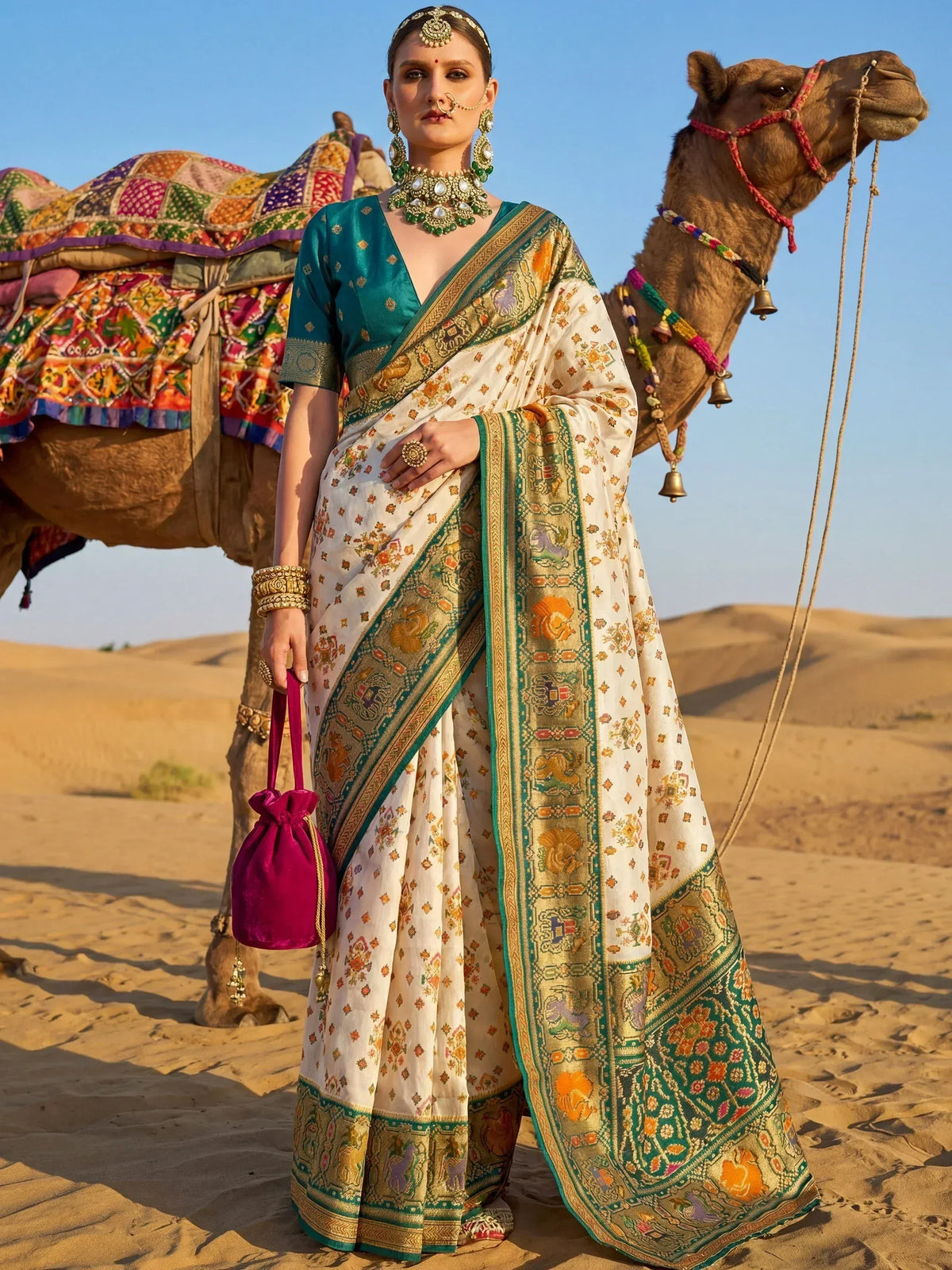 Woman in elegant white and green printed saree with temple border, standing beside camel in desert, holding pink potli bag
