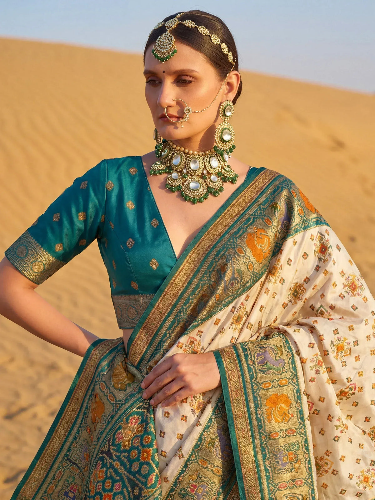 Elegant woman in teal and cream designer saree with gold embroidery, traditional Indian jewelry, desert backdrop
