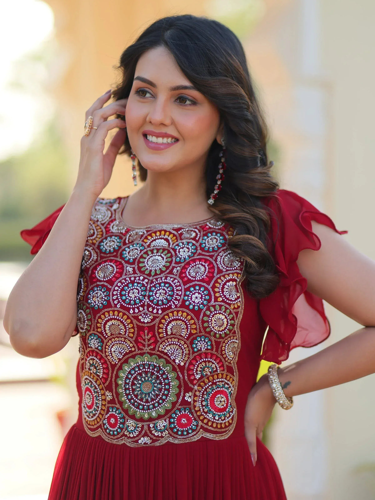 Woman in red embroidered ethnic gown with floral motifs, ruffled sleeves, and pleated skirt, styled with statement earrings.