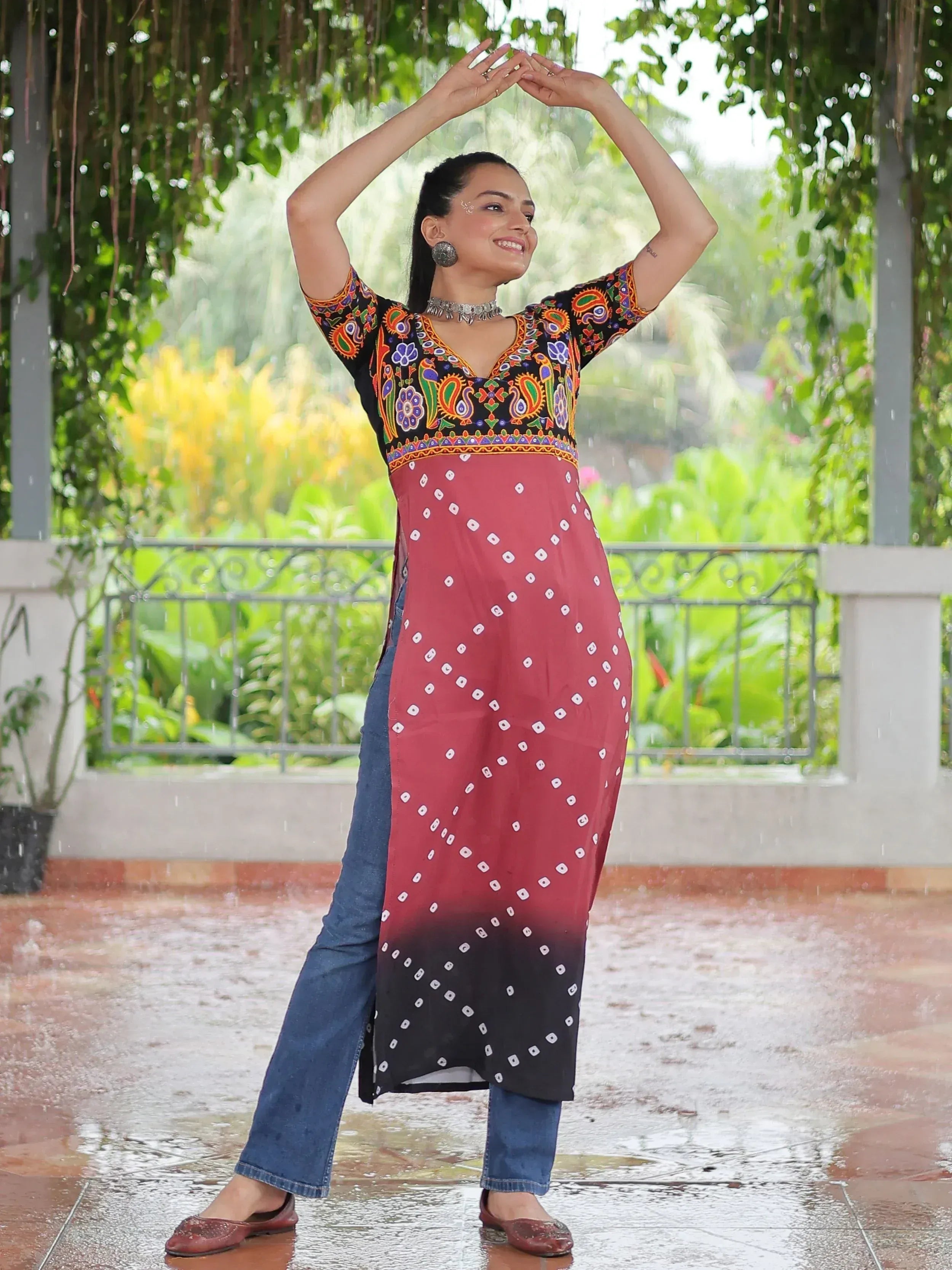 Woman in colorful embroidered kurta with bandhani print, paired with jeans, dancing in rain under greenery
