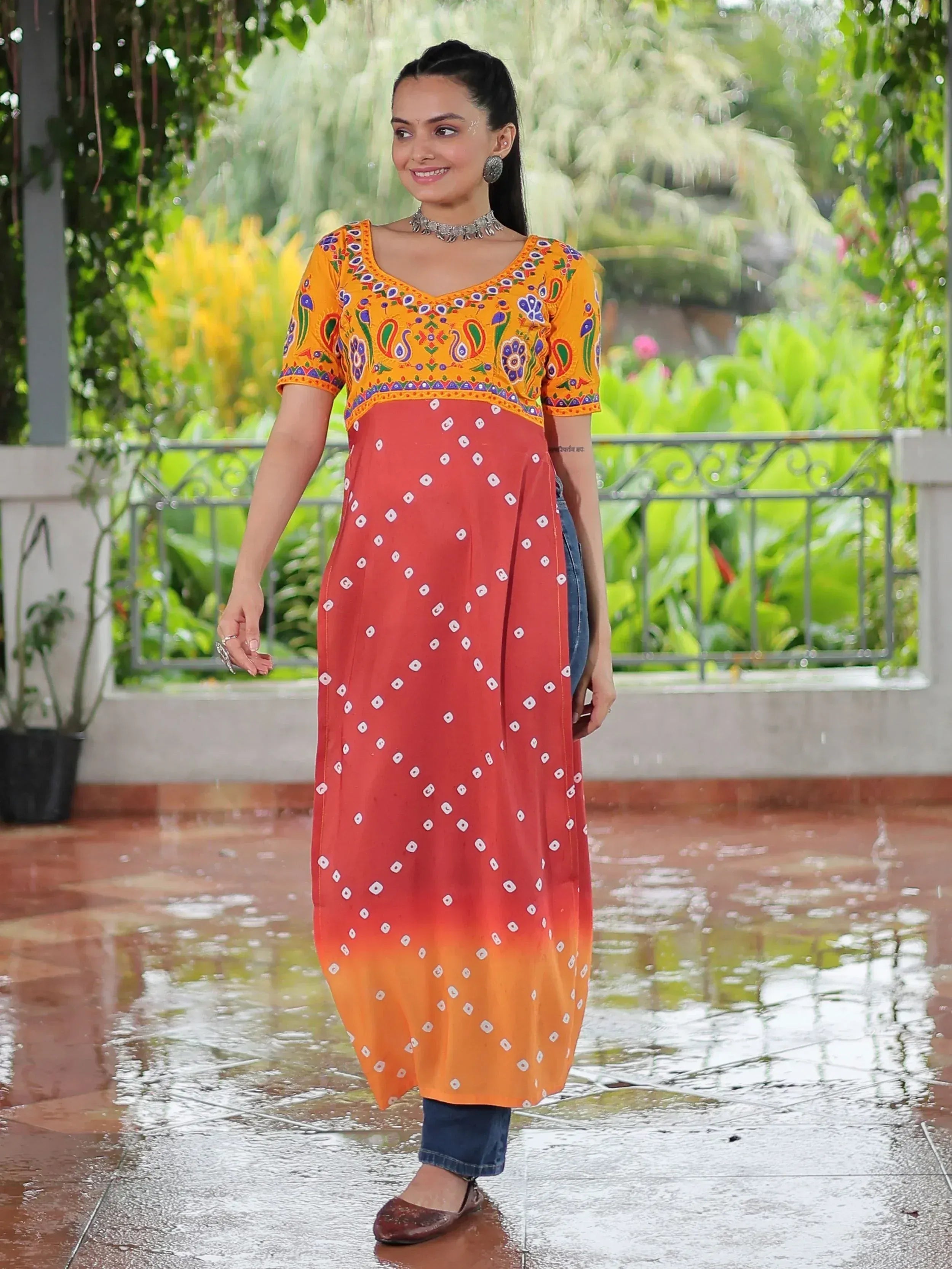 Woman in vibrant orange and red printed ethnic kurta with denim jeans, standing in rain on patio