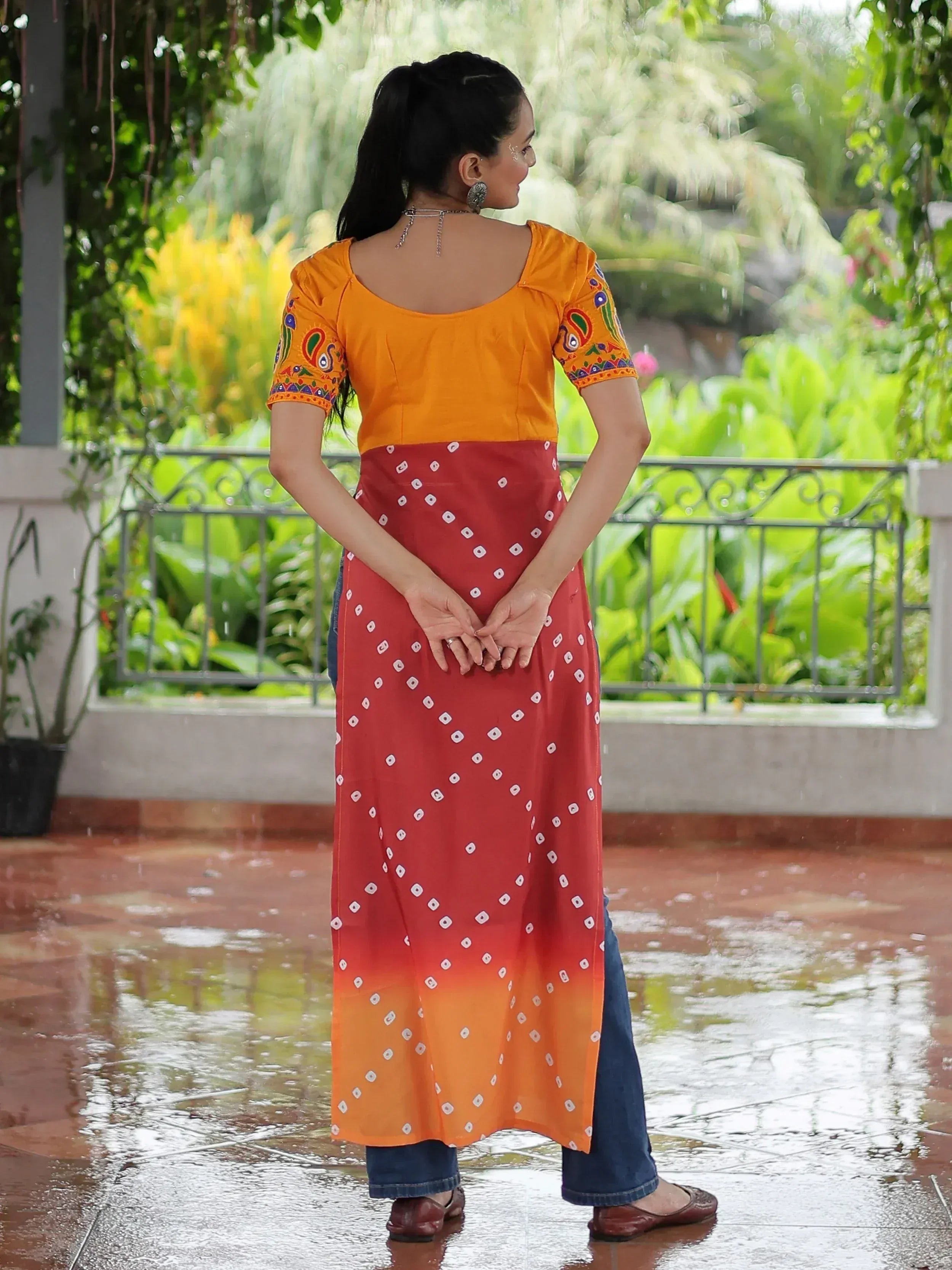 Woman in orange embroidered top and red bandhani print ethnic kurti with jeans, standing in rain