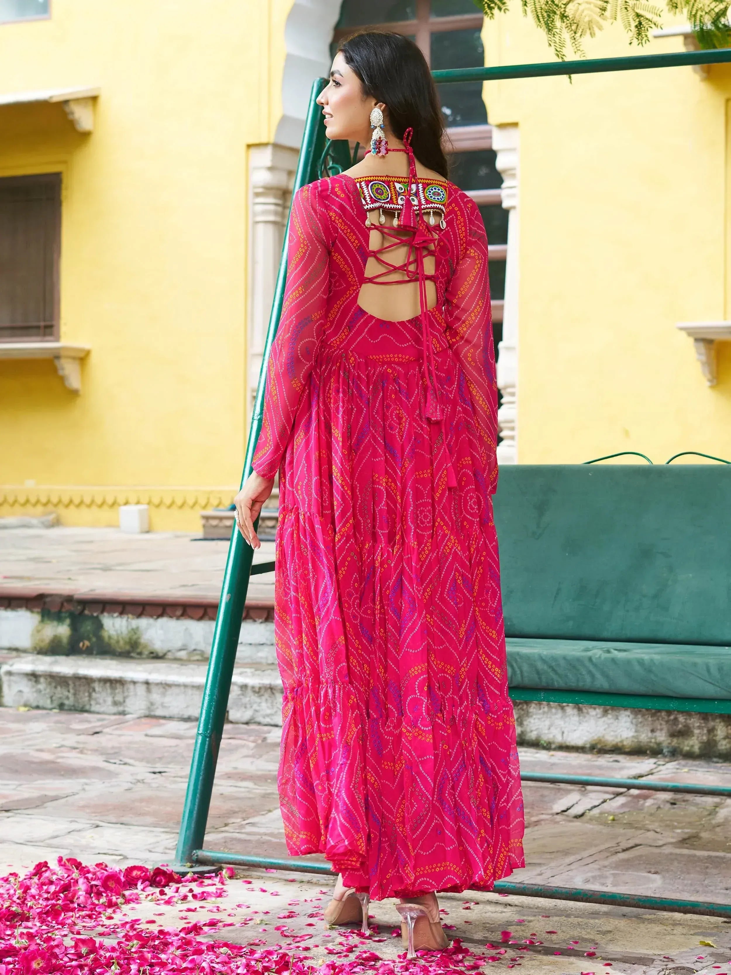 Woman in vibrant pink ethnic gown with back lace-up and mirror work, standing near yellow building with rose petals.