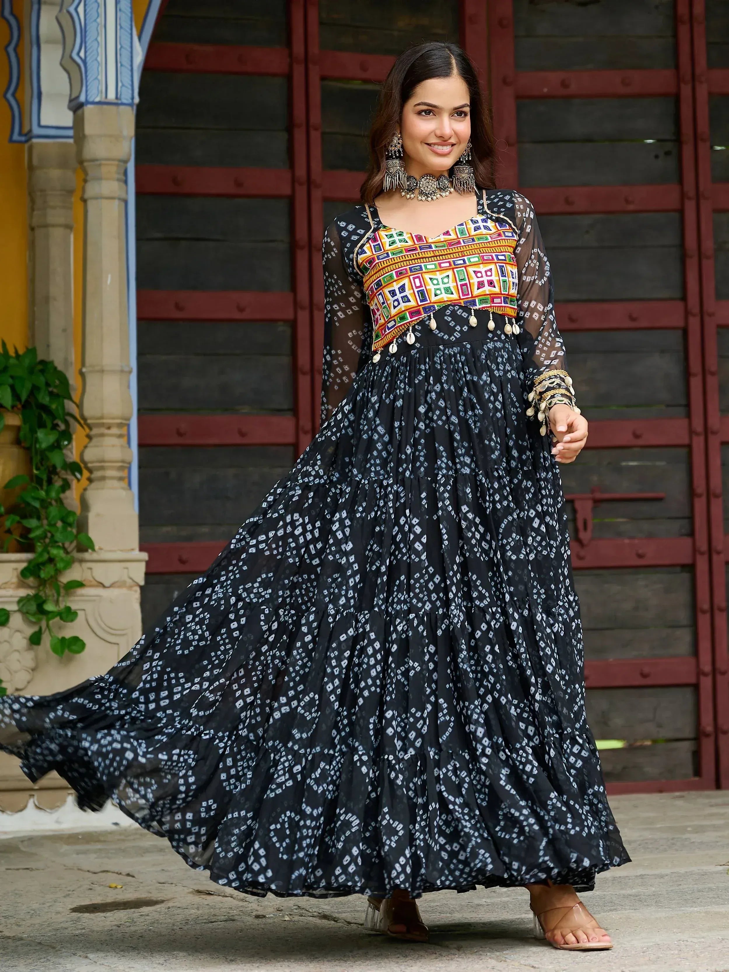 Woman in black and white printed ethnic gown with colorful embroidered top, traditional jewelry, and sheer sleeves.