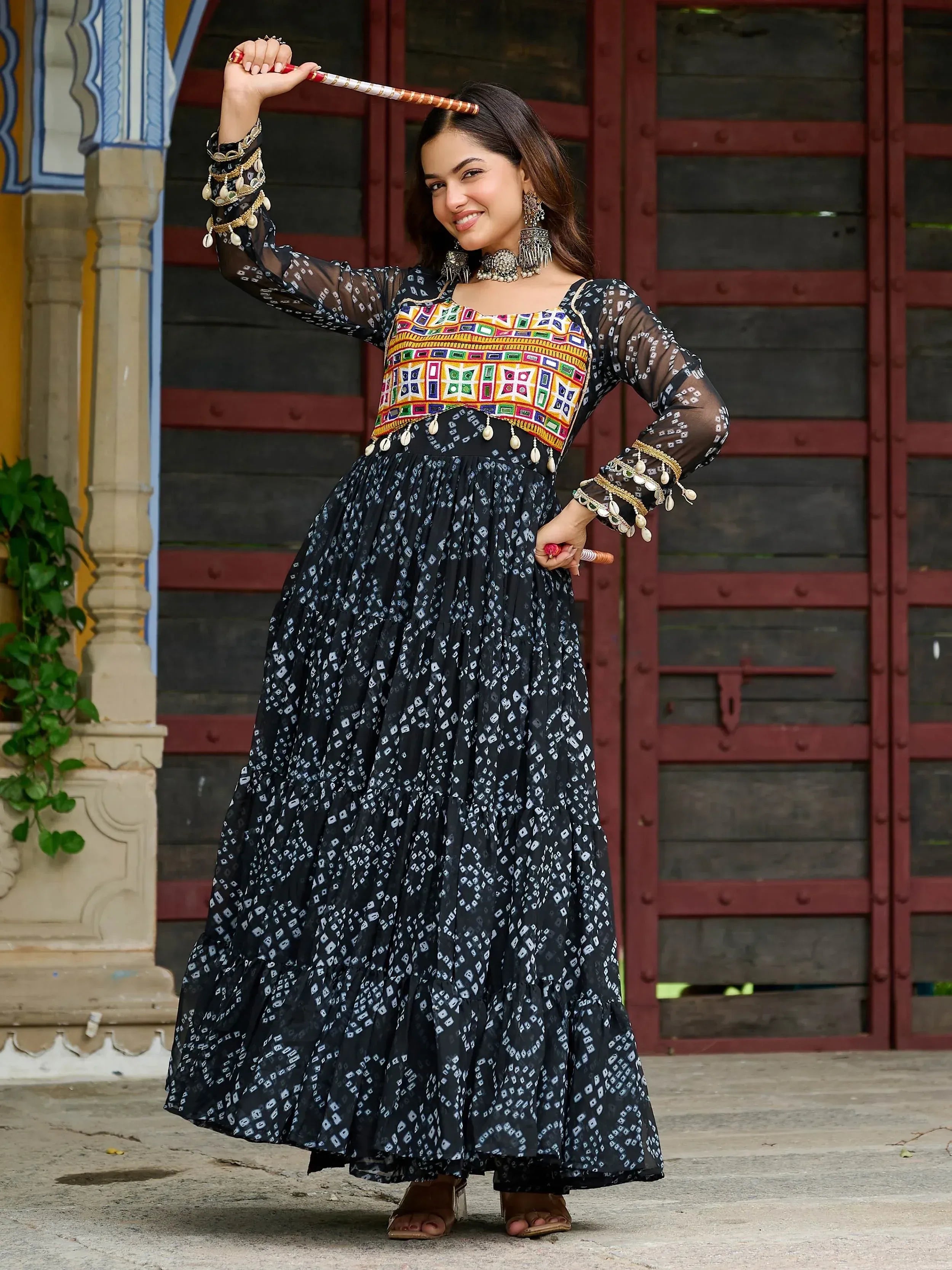 Woman in black printed ethnic gown with colorful embroidered bodice, holding decorative stick, traditional Indian wear