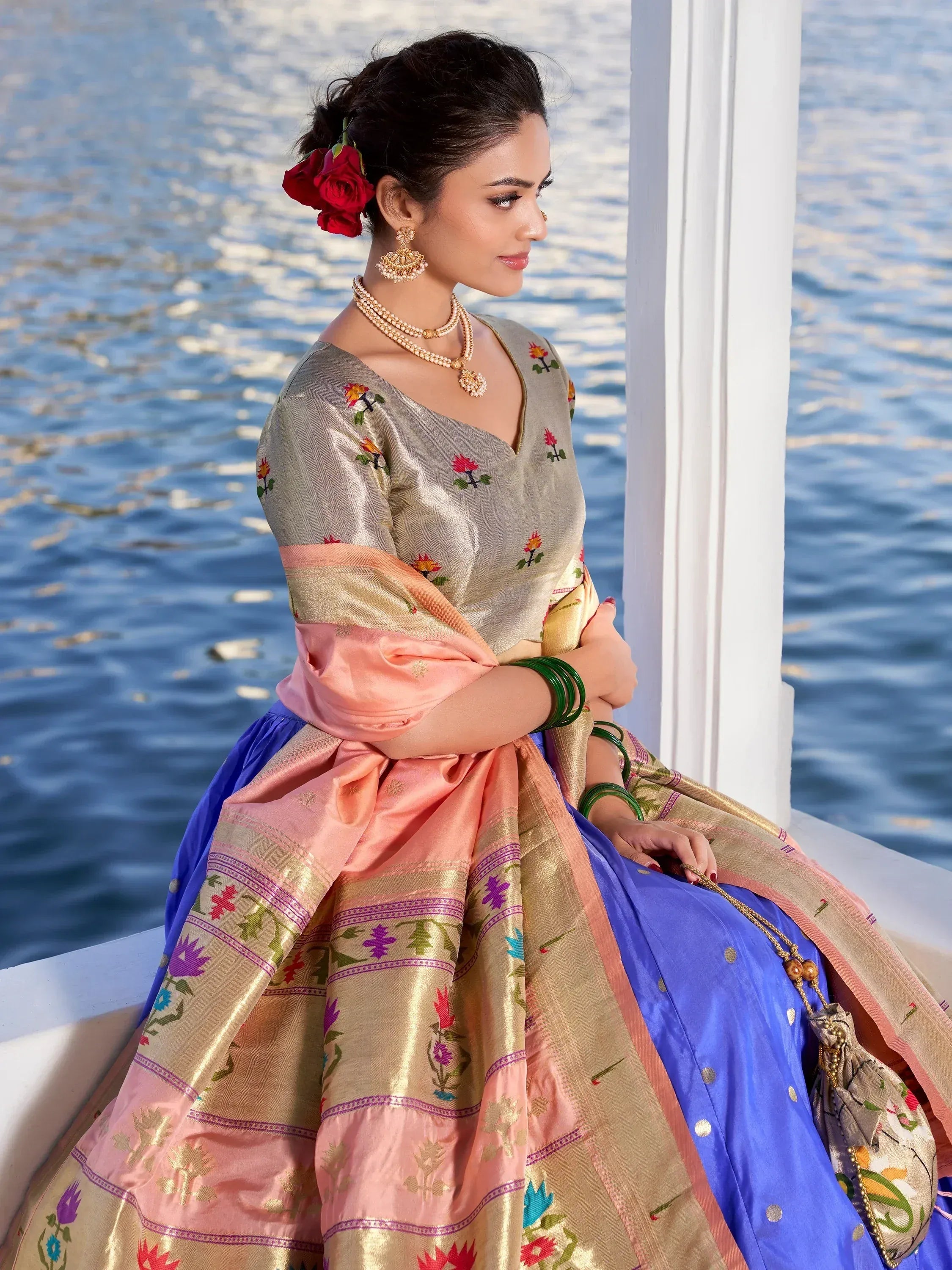 Woman in elegant Indian ethnic saree with floral embroidery, gold border, and blue skirt, accessorized with traditional jewelry by water