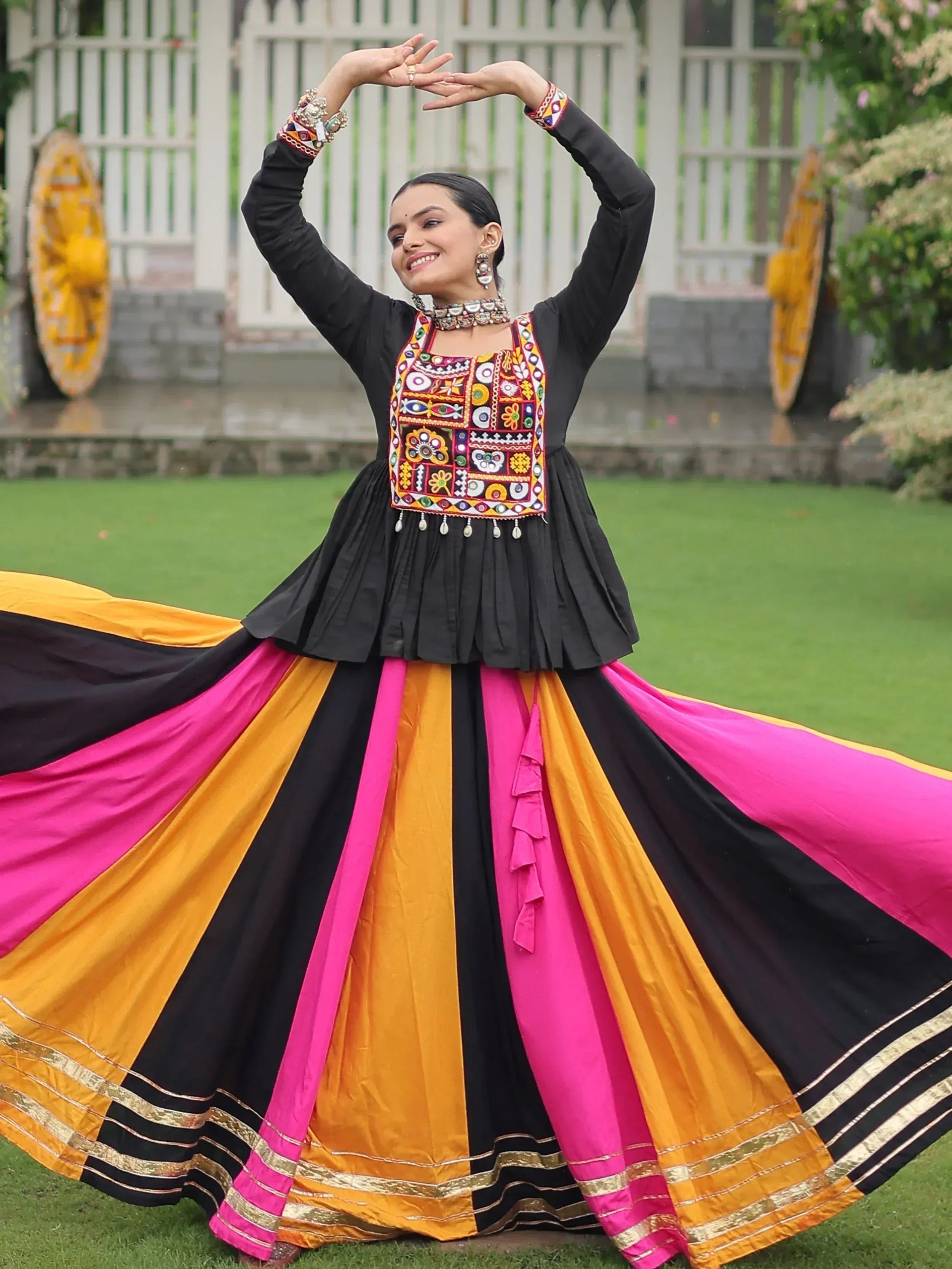 Woman in vibrant black, pink, and orange ethnic gown with mirror work, dancing outdoors.