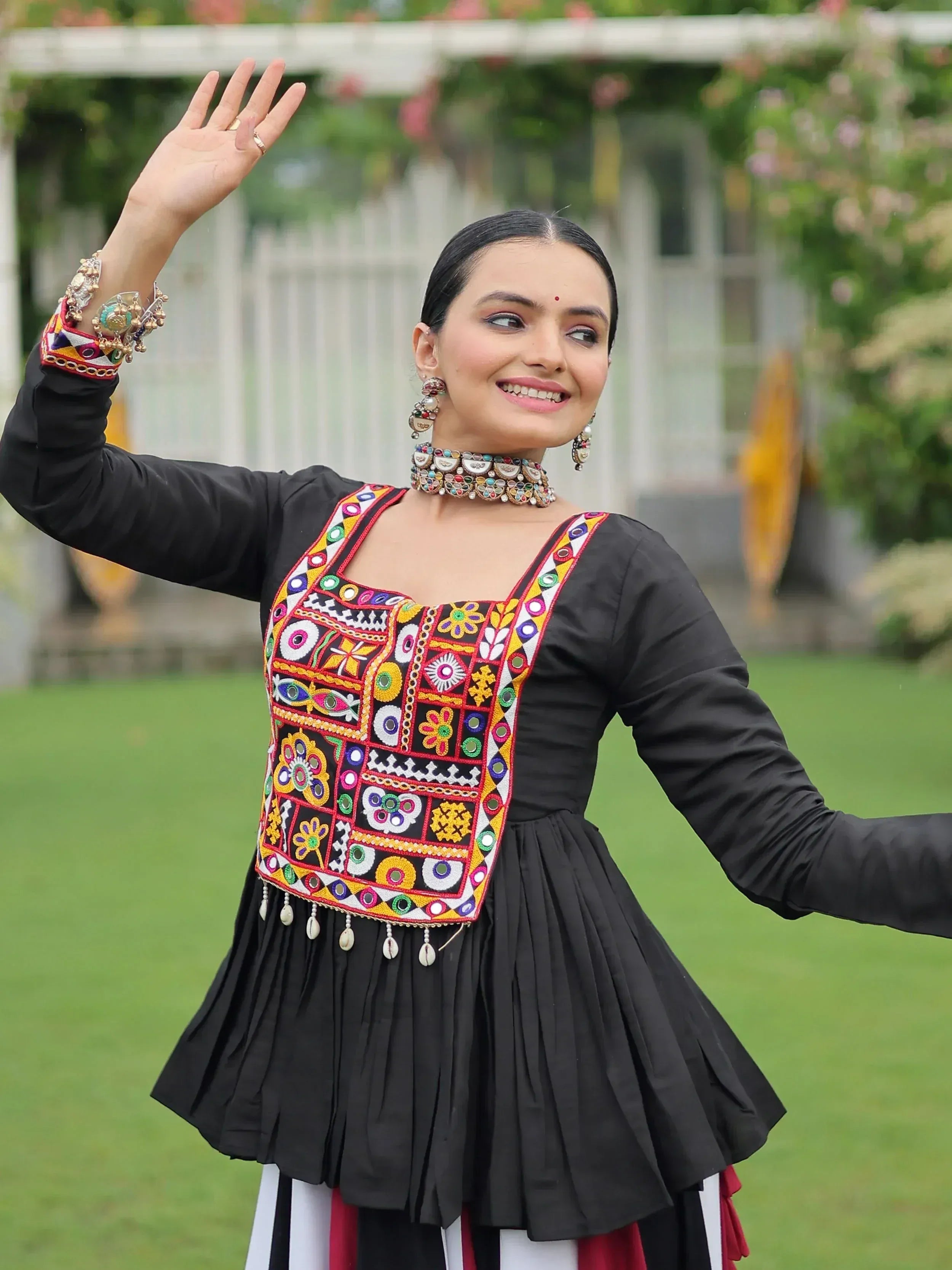 Woman in black ethnic dress with colorful mirror embroidery and traditional jewelry, waving outdoors.