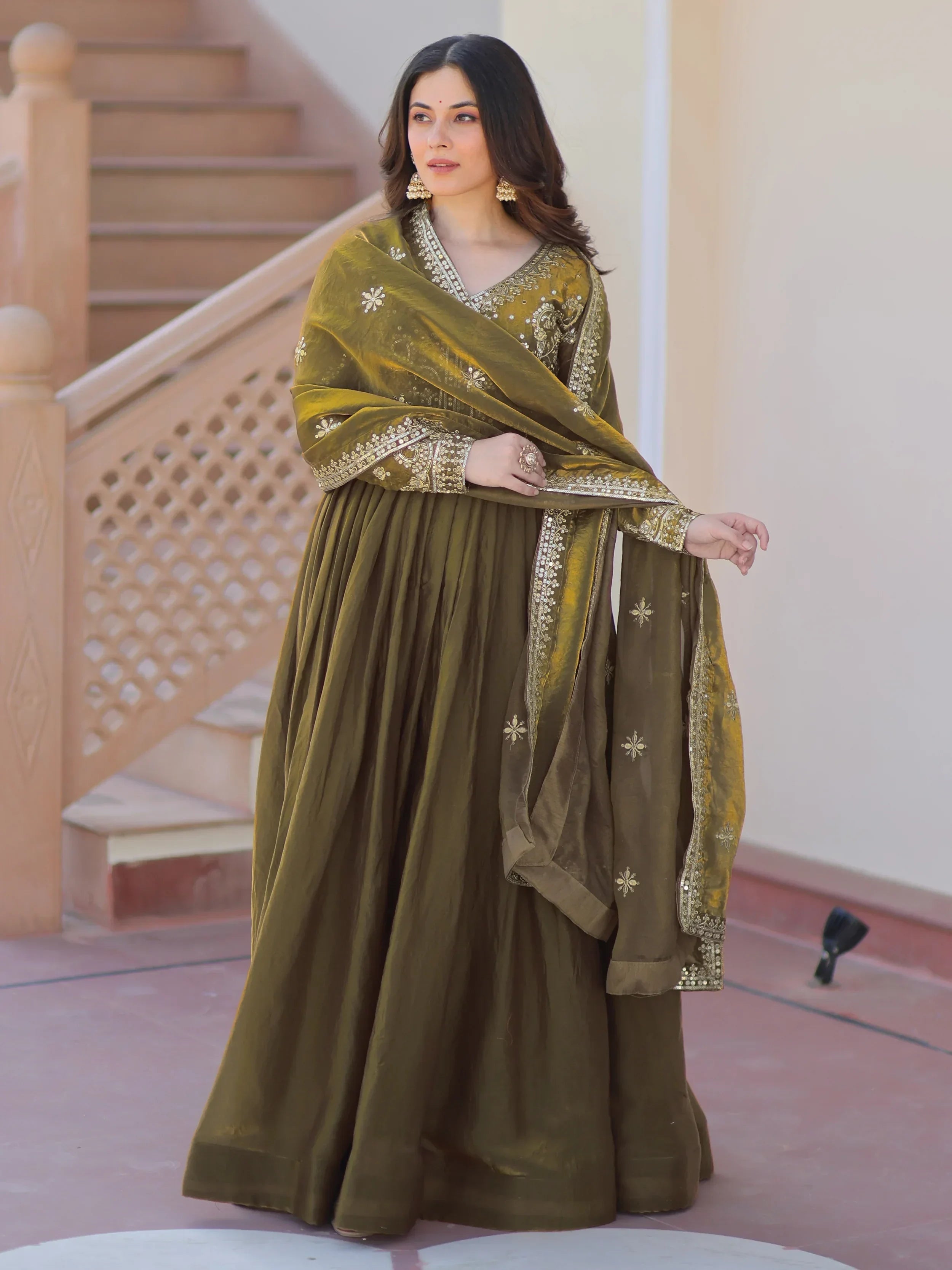 Woman in olive green embroidered ethnic gown with dupatta, standing outdoors.
