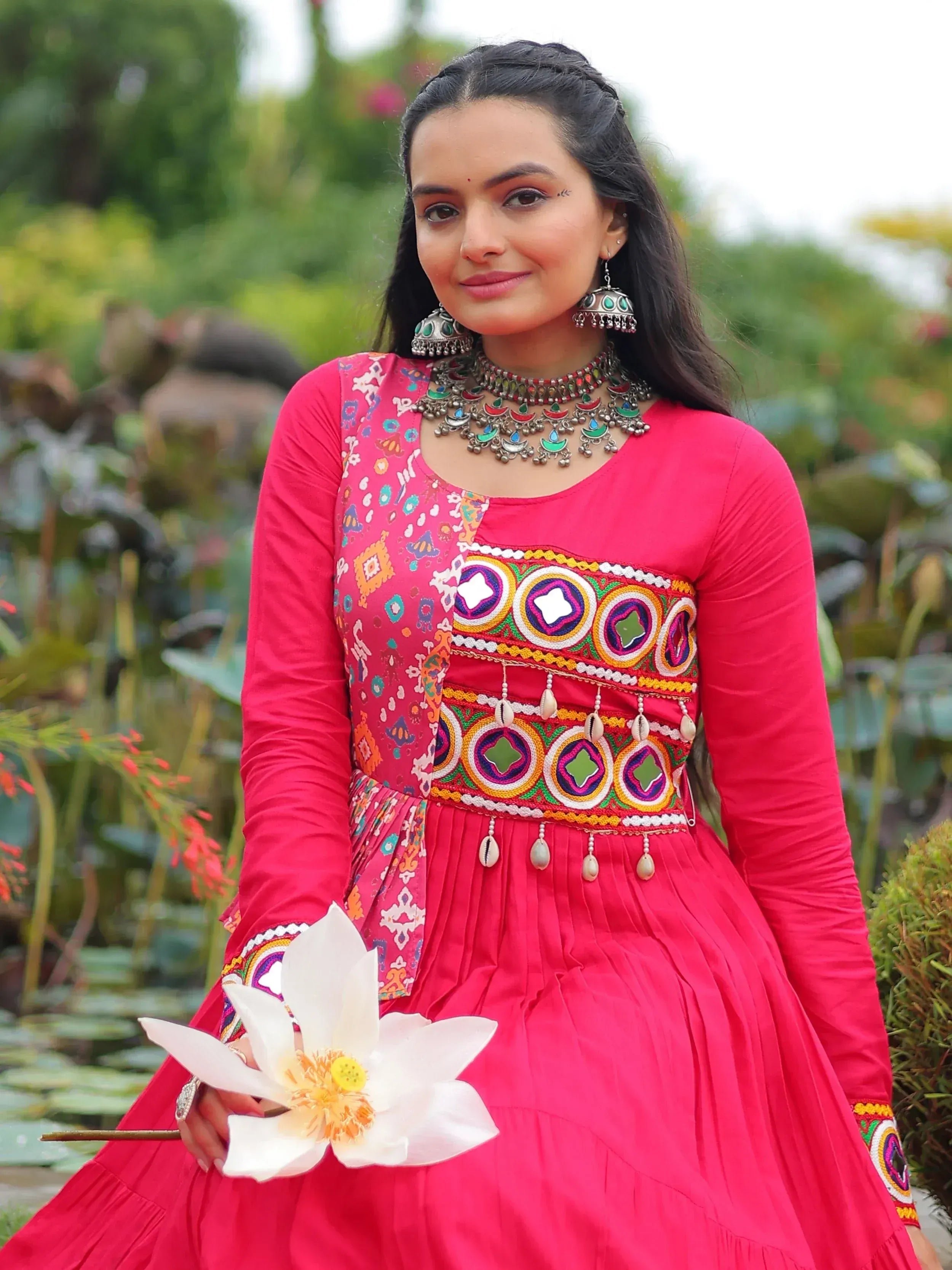 Woman in vibrant pink embroidered ethnic gown with traditional jewelry, holding white lotus flower outdoors