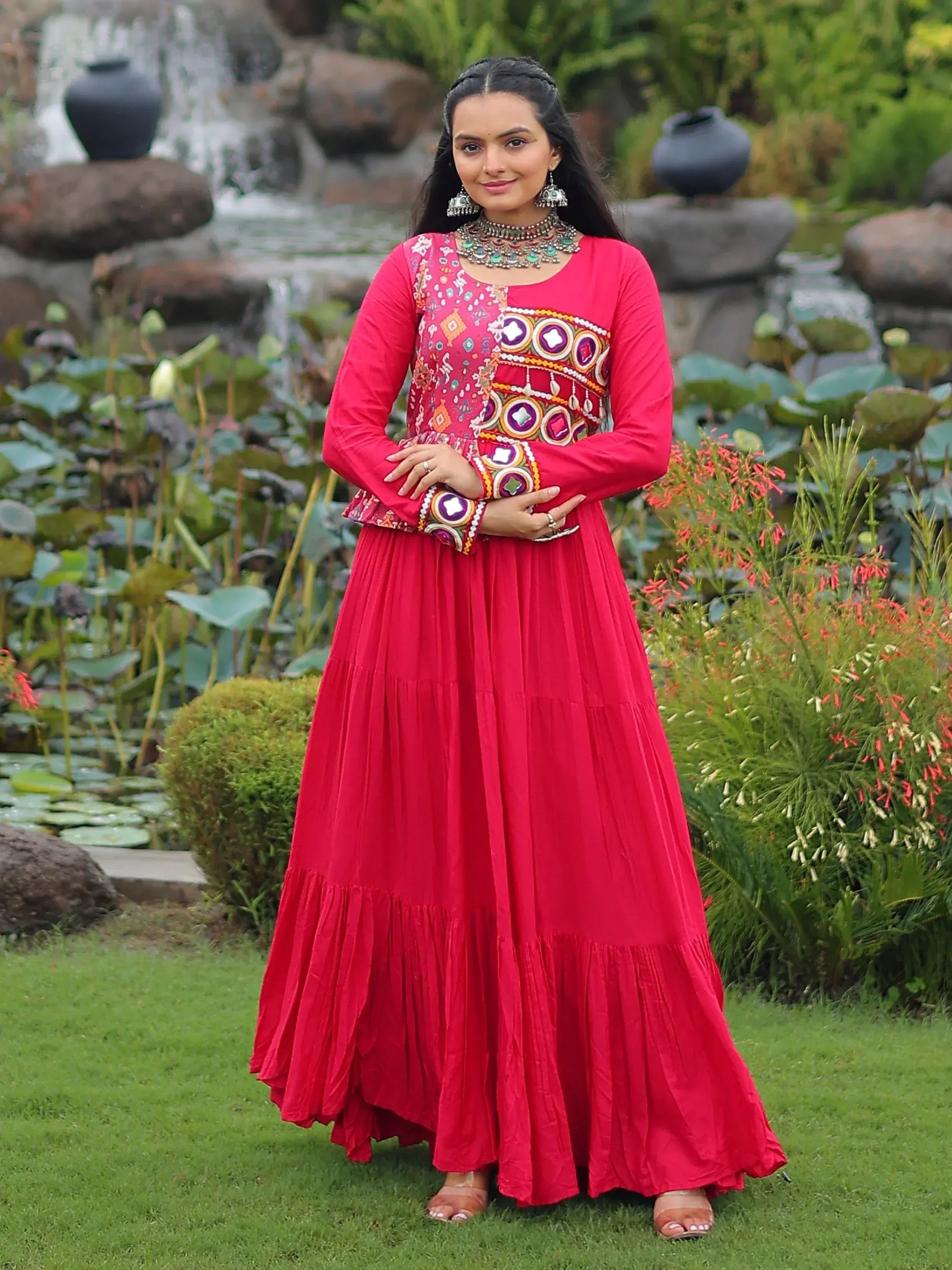 Woman in vibrant red ethnic gown with mirror embroidery, standing in garden near waterfall