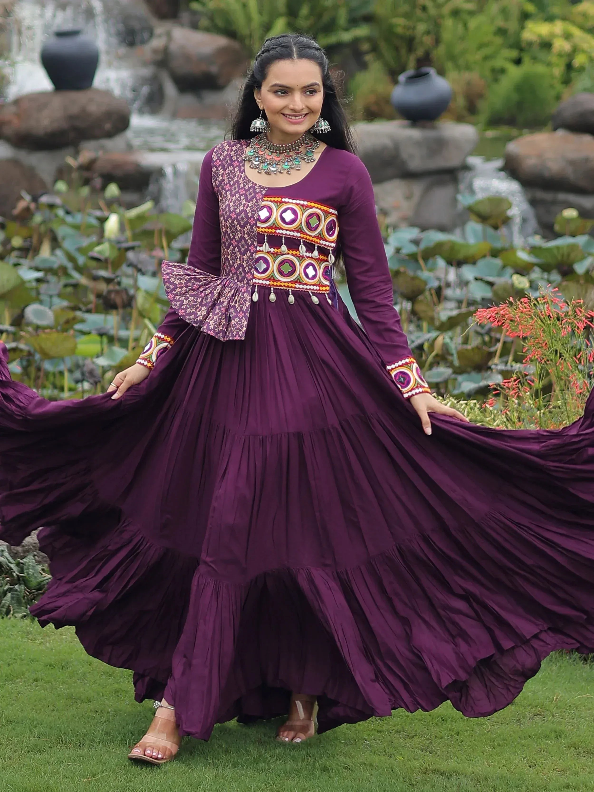 Woman in purple ethnic gown with mirror work, pleated skirt, and traditional jewelry, standing in garden