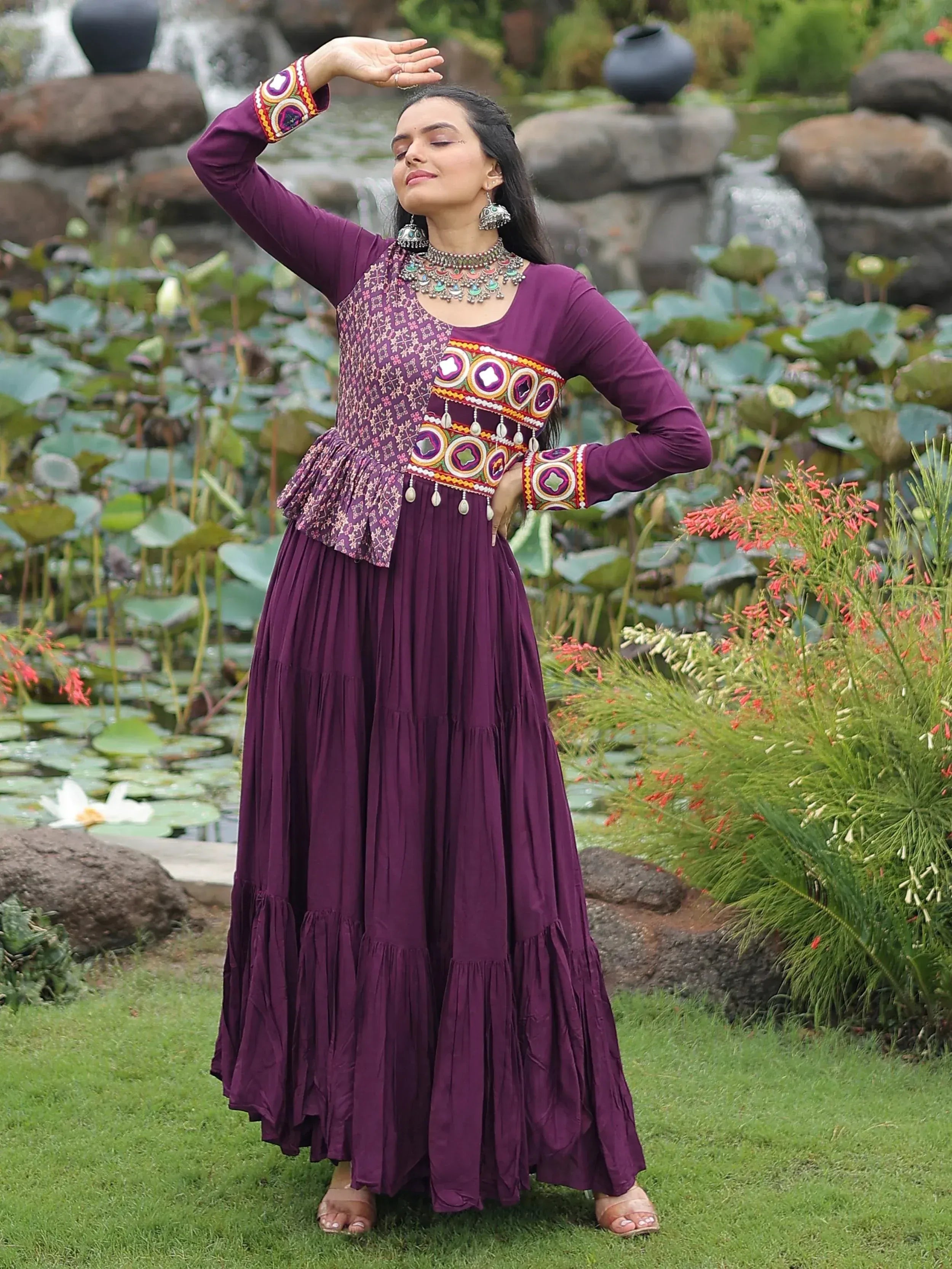 Woman in purple ethnic gown with mirror work, standing by garden pond, perfect for festive occasions