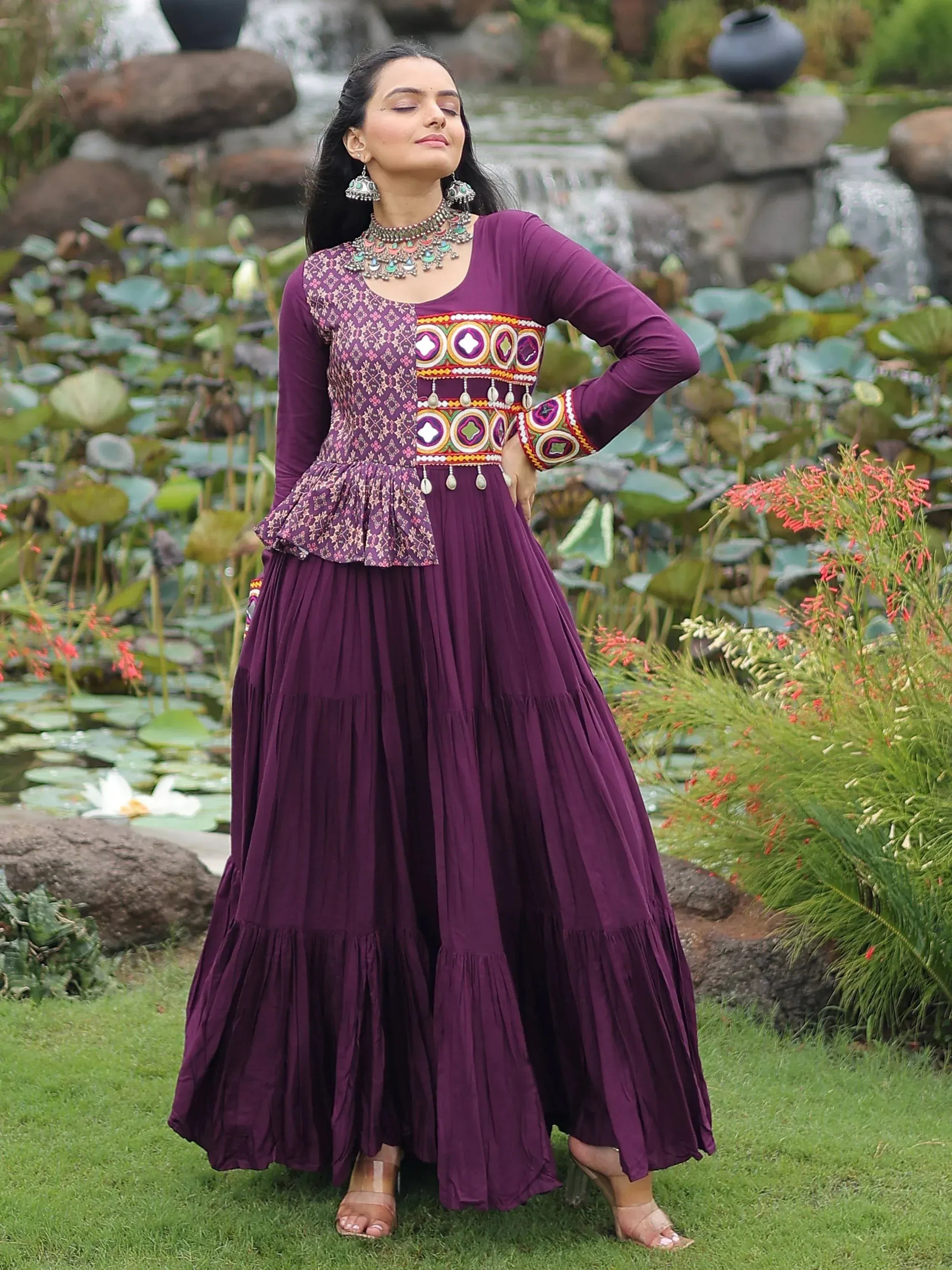 Purple ethnic gown with embroidered peplum top and pleated skirt, styled with traditional jewelry in garden setting
