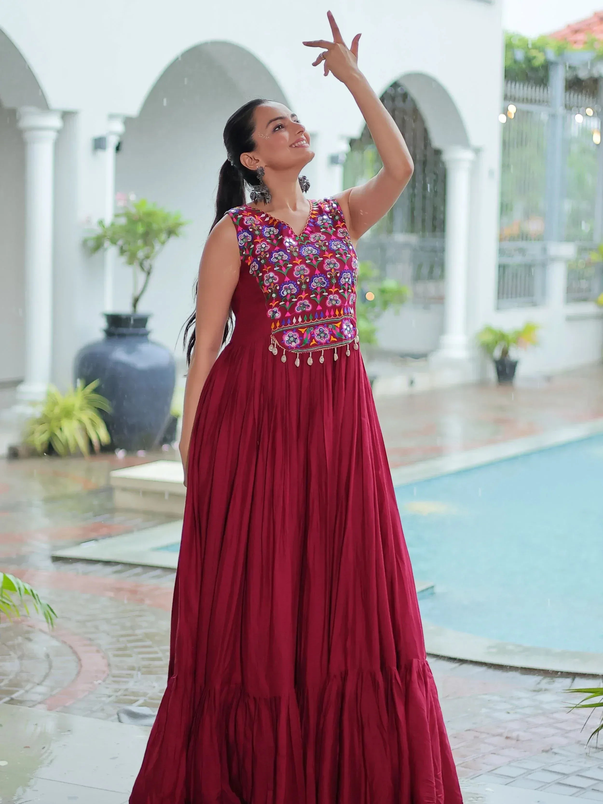 Woman in maroon ethnic gown with floral embroidery, standing by pool in rain