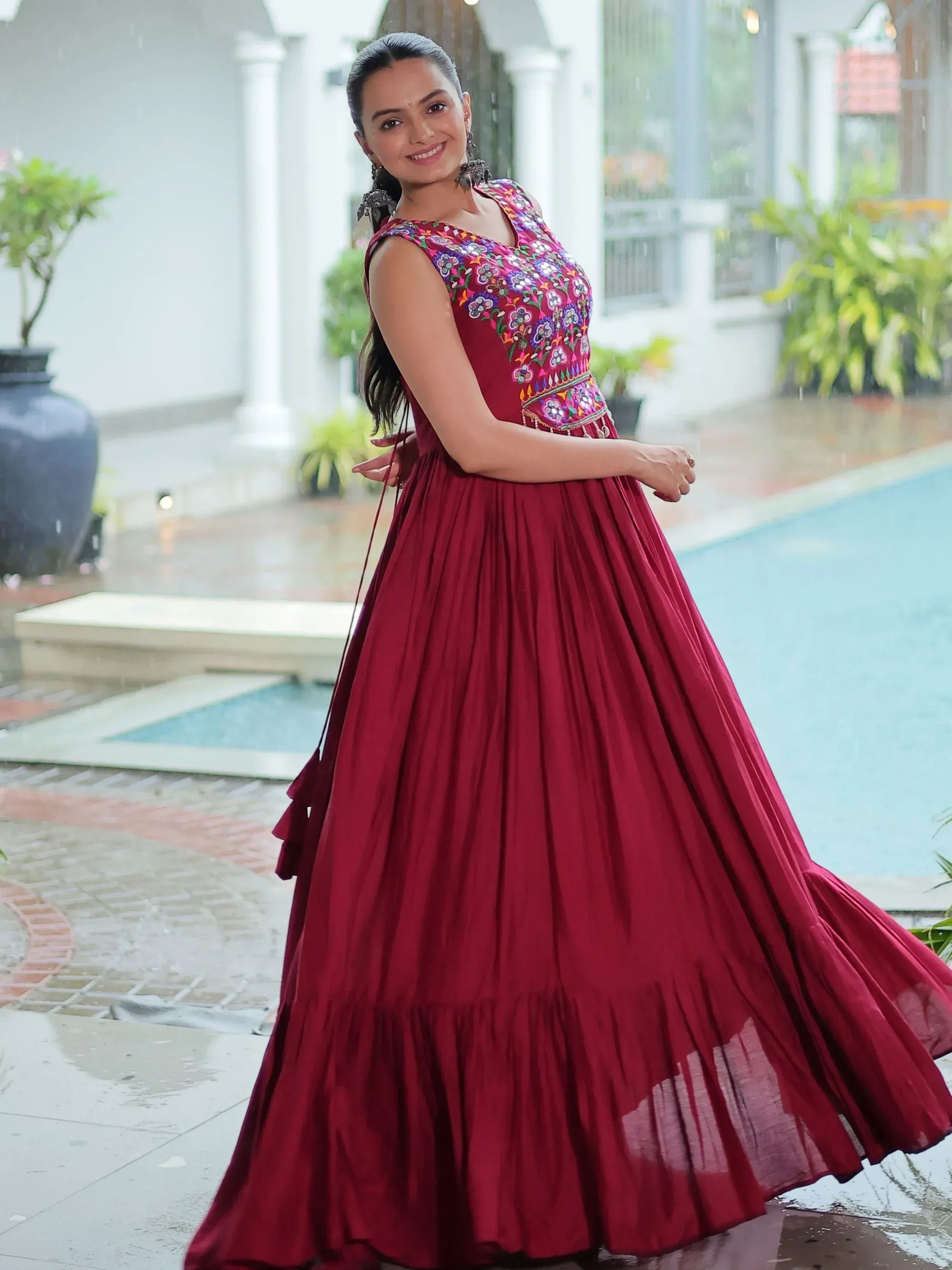 Woman in maroon embroidered ethnic gown with floral motifs, standing by poolside in rain